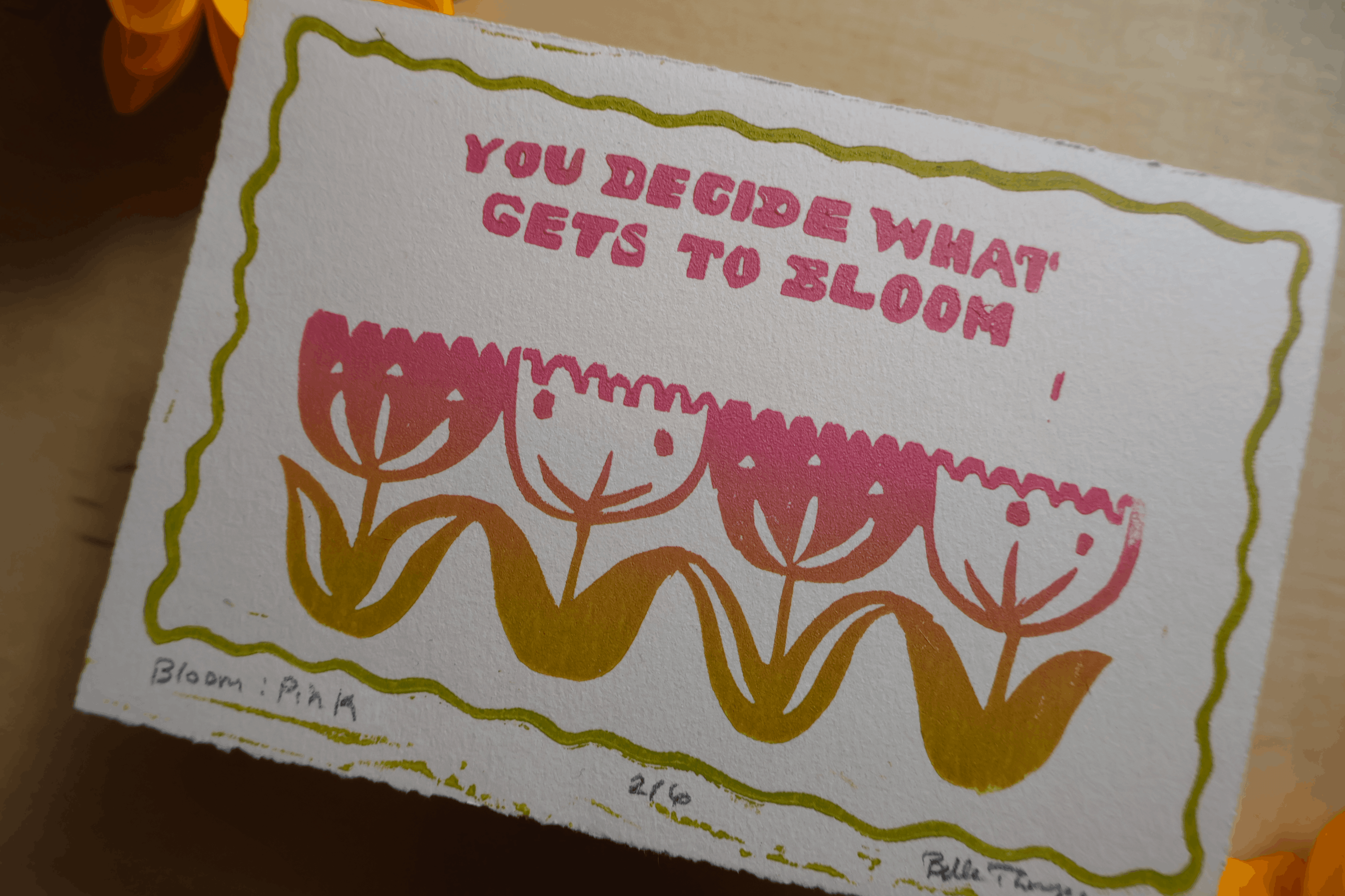 A square white art print or card with a hand-drawn olive green border, pink tulip illustration, and the phrase 'YOU DECIDE WHAT GETS TO BLOOM' in pink text, photographed on a warm wooden surface.