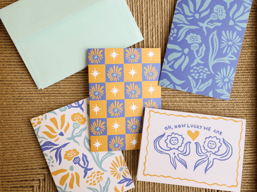 Four decorative cards and an envelope arranged on a woven brown surface. The set includes a pale mint envelope, a yellow-and-blue floral checkerboard card, a blue-and-white floral card, a white card with blue floral artwork and the text 'Oh, how lucky we are,' and another floral card partially visible underneath.