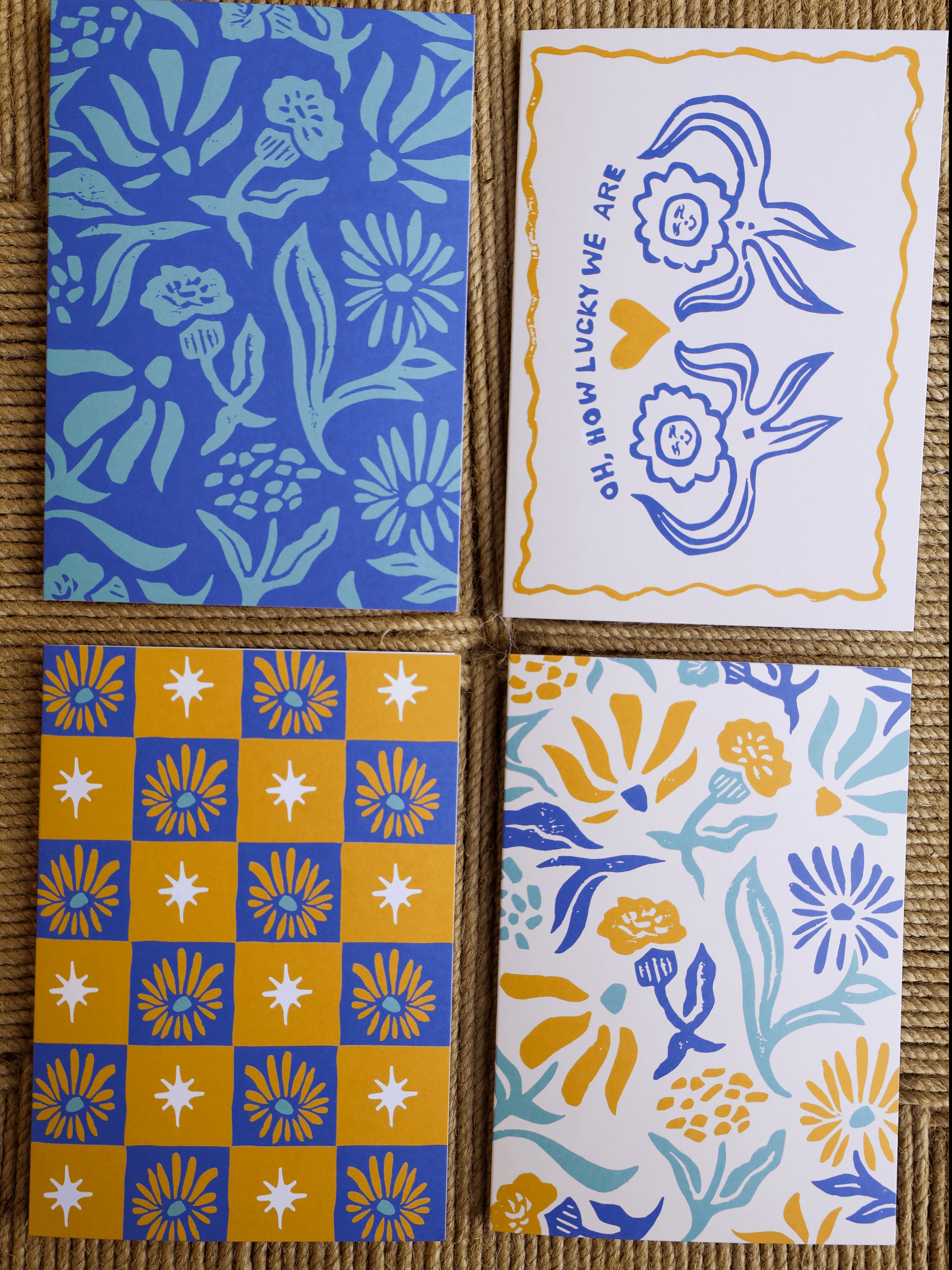 Four square greeting cards arranged in a 2x2 grid on a brown woven mat background. The cards feature blue, yellow, and light blue floral and geometric patterns, including one card with blue bunny illustrations and the text 'GIFT HOW LUCKY WE ARE'.
