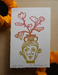 An art print featuring a stylized face in a vase with red flowers, photographed flat on a light wood surface with sunflower petals around it.