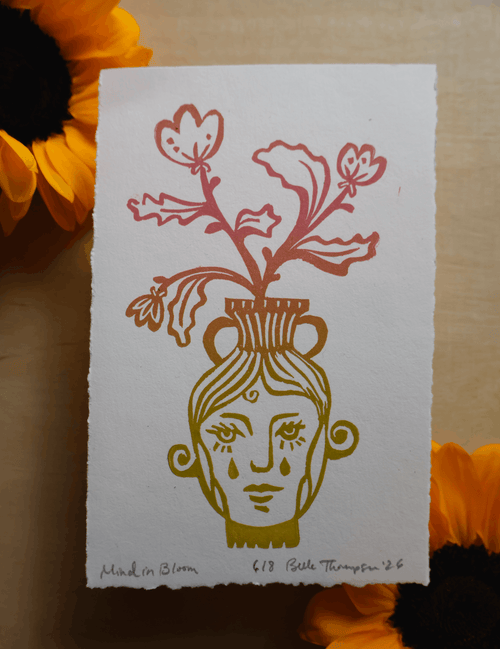 An art print featuring a stylized face in a vase with red flowers, photographed flat on a light wood surface with sunflower petals around it.