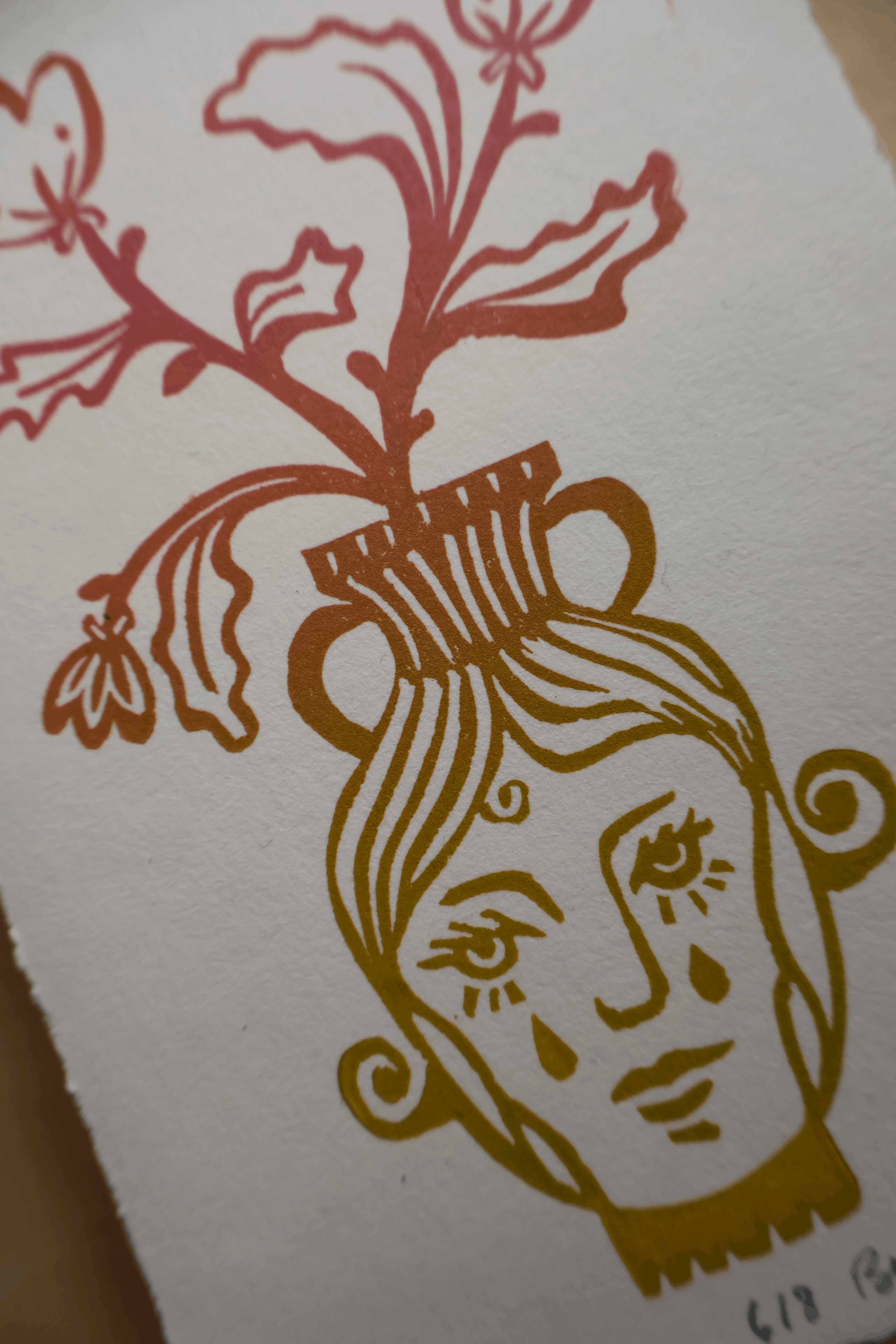 A close-up of a textured white paper print featuring a stylized illustration of a face with a floral pot on its head, rendered in warm gold and muted red ink.