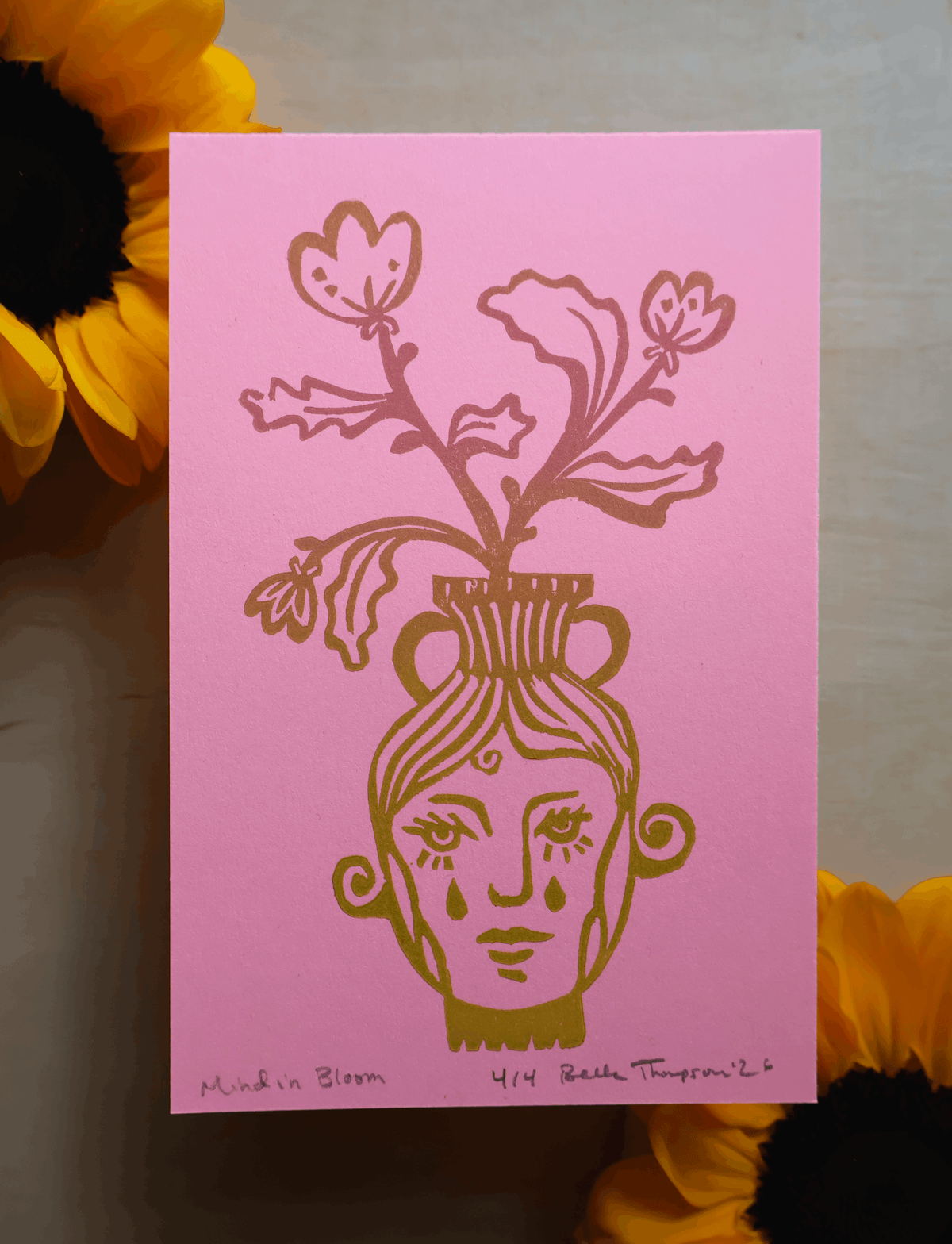A pink art print featuring a stylized face with tears flowing from a vase of flowers, styled with yellow sunflowers in the corners.