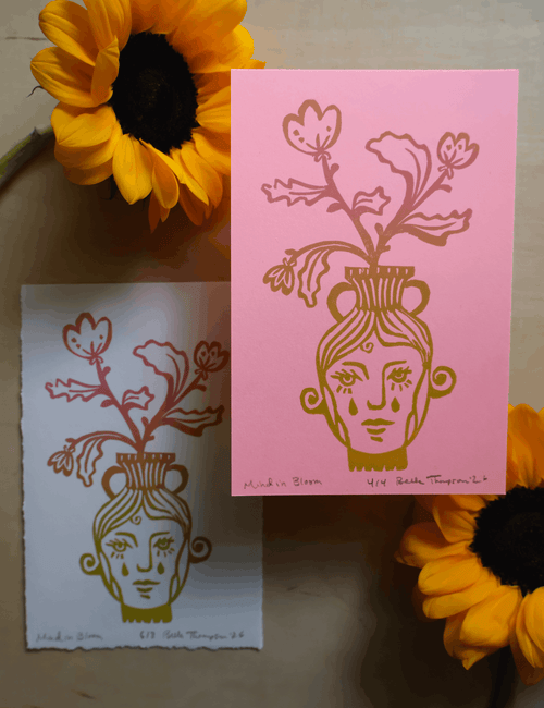 Two art prints featuring a stylized face in a vase with flowers, styled with yellow sunflowers on a wooden surface. One print is pink on white and the other is white on pink, with handwritten text at the bottom.