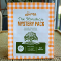 florida themed mystery pack packaging showing the dovetail logo deer and willow tree print design and contents of a print, stickers, and sew on patch with a blurred background