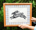 A framed art print featuring a black ink illustration of a rabbit with floral patterns, held by hands against a background of green foliage. The print has a patterned border and is housed in a light brown wooden frame.