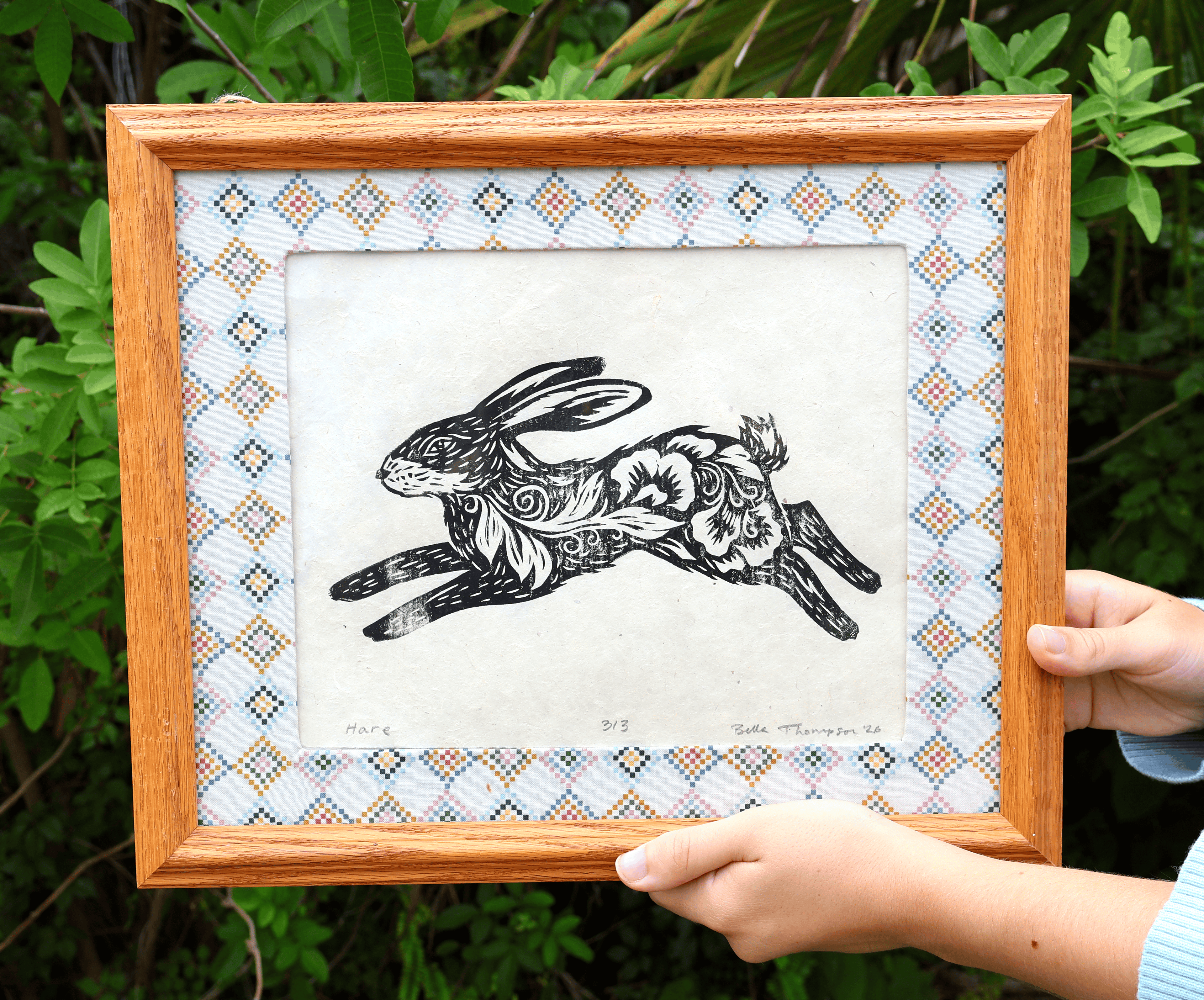 A framed art print featuring a black ink illustration of a rabbit with floral patterns, held by hands against a background of green foliage. The print has a patterned border and is housed in a light brown wooden frame.