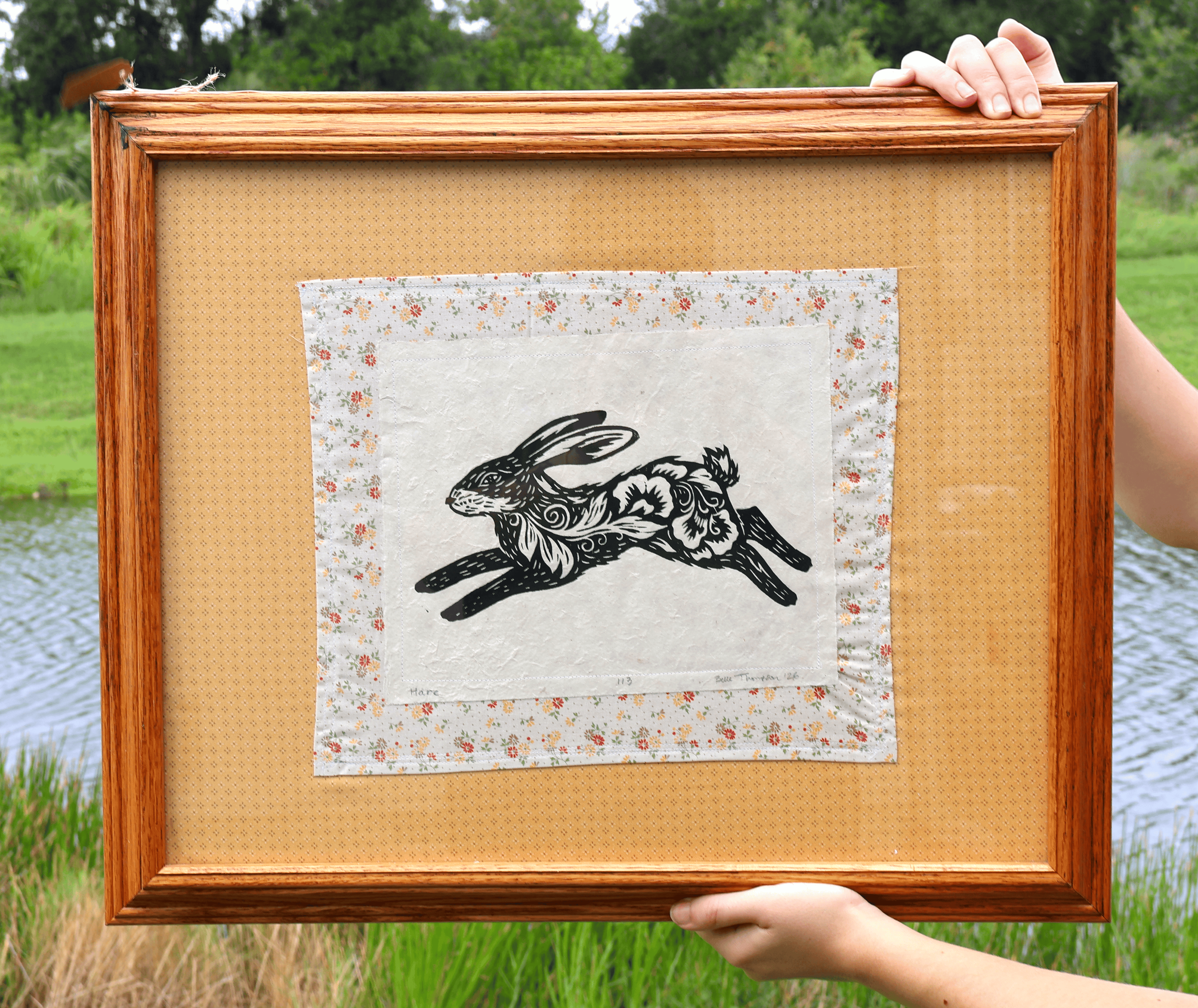 A person holds a square framed art print featuring a black rabbit illustration on a cream fabric background with a floral border, set against an outdoor pond and grass backdrop.