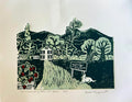 full color with light green, dark green, cream, and red handmade linocut print showing a strawberry farm landscape with a house, trees, a box of strawberries, and a dog on a piece of heavyweight paper held by a hand with a blurred background