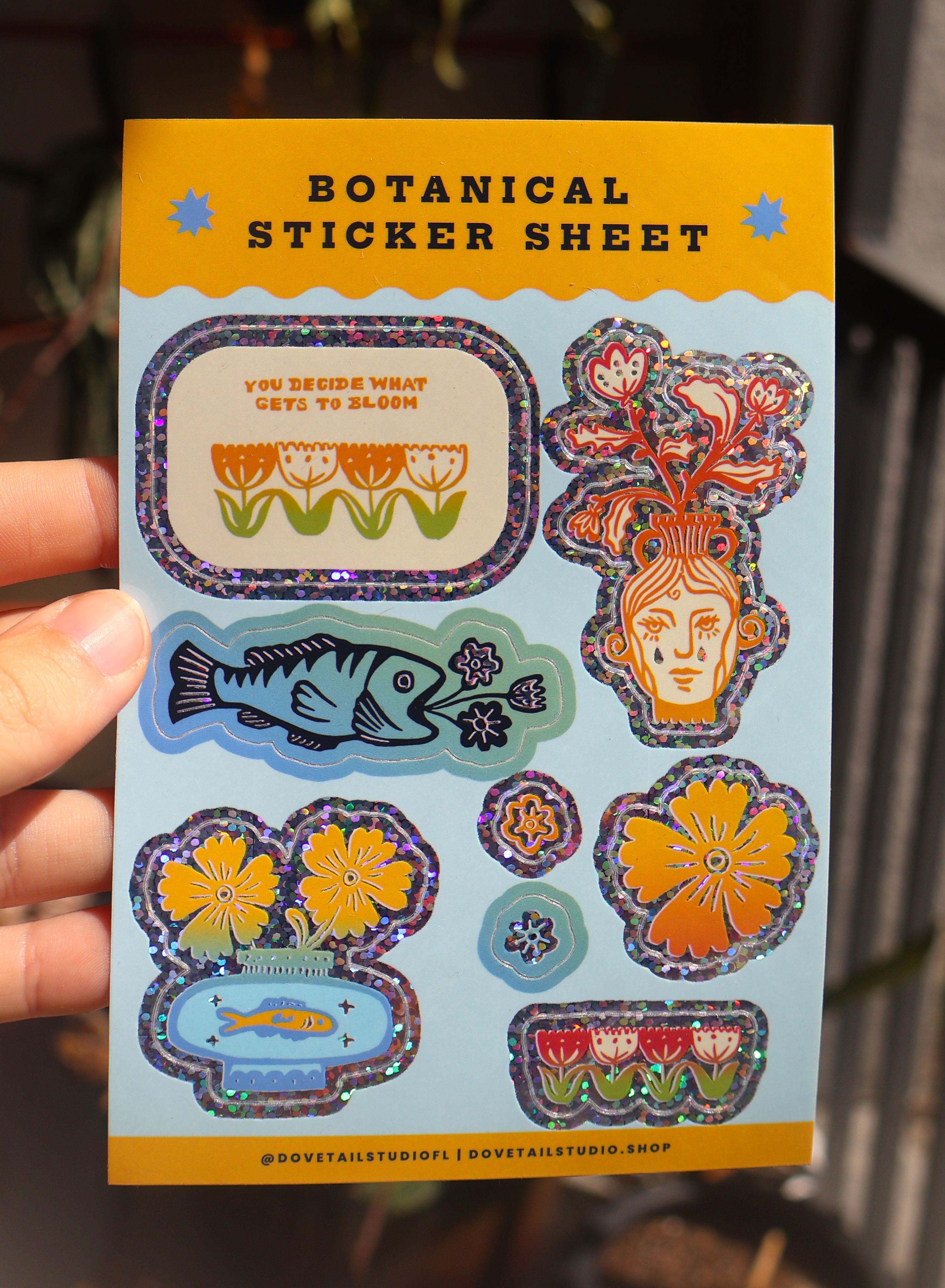 A hand holds a botanical sticker sheet featuring colorful illustrated flowers, fish, and a stylized face with floral elements, on a light blue backing card with yellow borders.