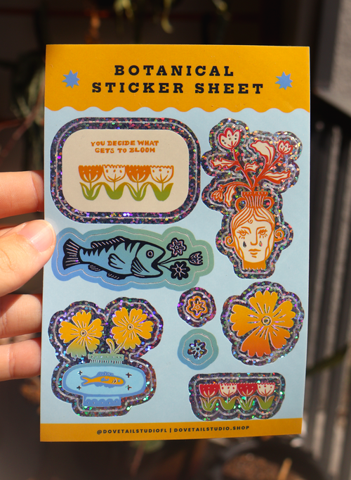 A hand holds a botanical sticker sheet featuring colorful illustrated flowers, fish, and a stylized face with floral elements, on a light blue backing card with yellow borders.