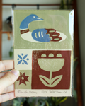 heron handmade linocut print in the colors blue brown and green on colored heavyweight paper signed by artist held by hand on blurred background