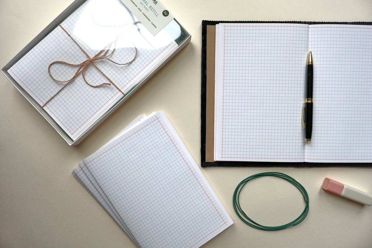 A styled flat-lay of stationery items on a light beige surface: an open navy-bound grid paper journal with a black and gold pen, a boxed pack of grid paper tied with twine, several loose grid paper sheets, a pink-and-white highlighter, and a dark green loop of elastic journal binders. showcasing dovetail studio's handmade refillable journal refills
