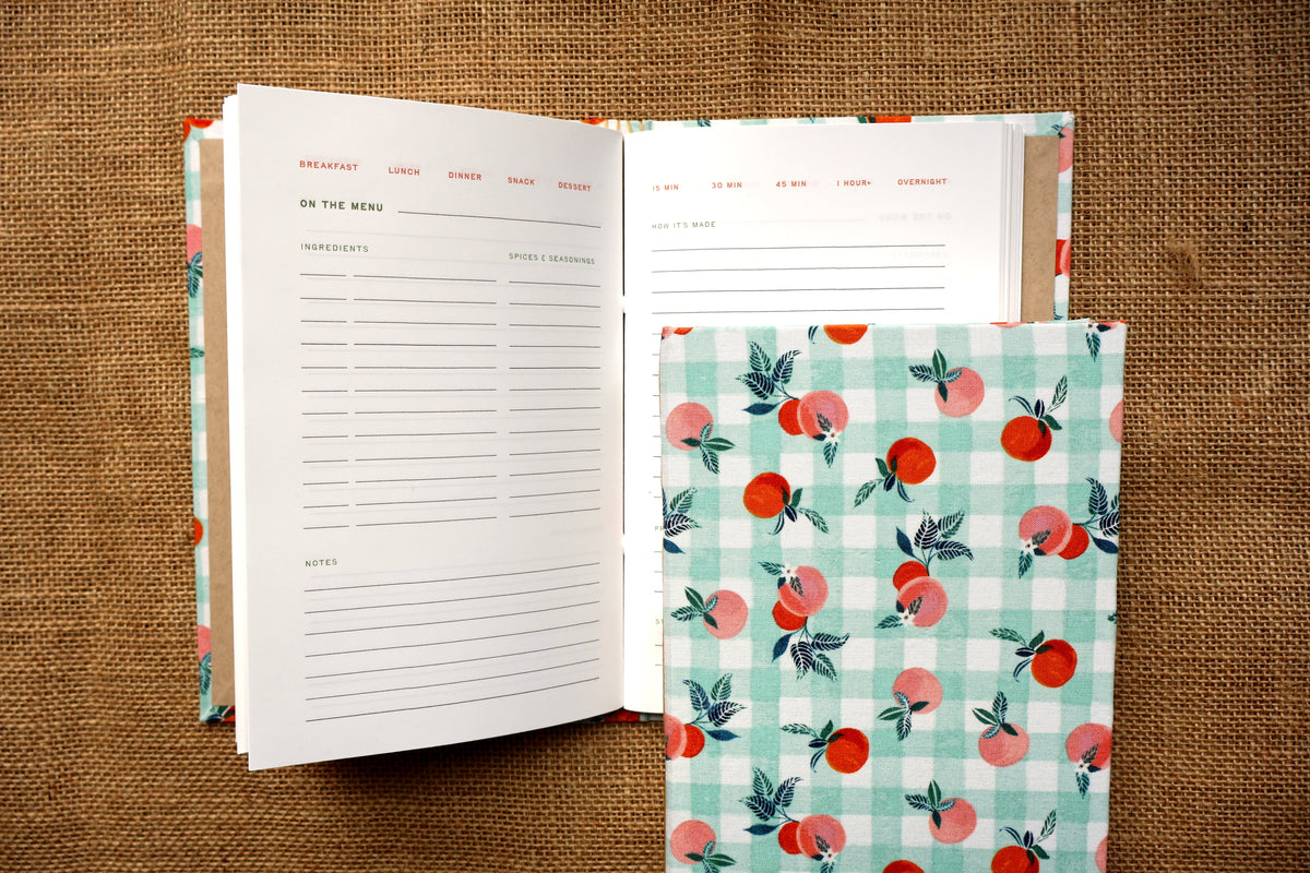 A fabric recipe log with a peach and gingham pattern, shown laid over an open lined recipe log on a burlap surface, with a cucumber and whisk as props. Handmade original design