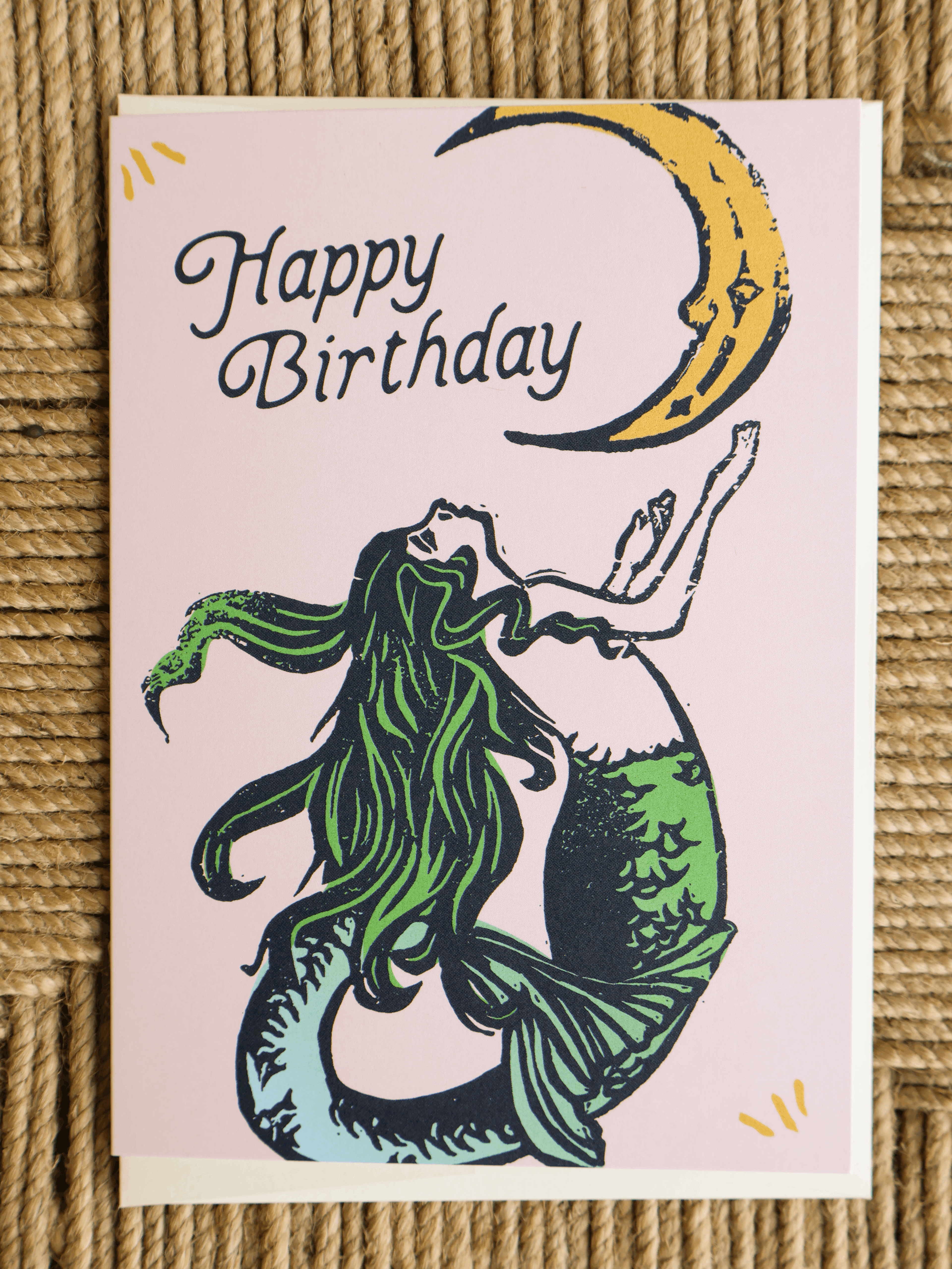 A square birthday card with a pink background, featuring a mermaid illustration with green hair and tail, a yellow crescent moon, and the words 'Happy Birthday' in black script. The card is photographed flat on a woven straw surface.