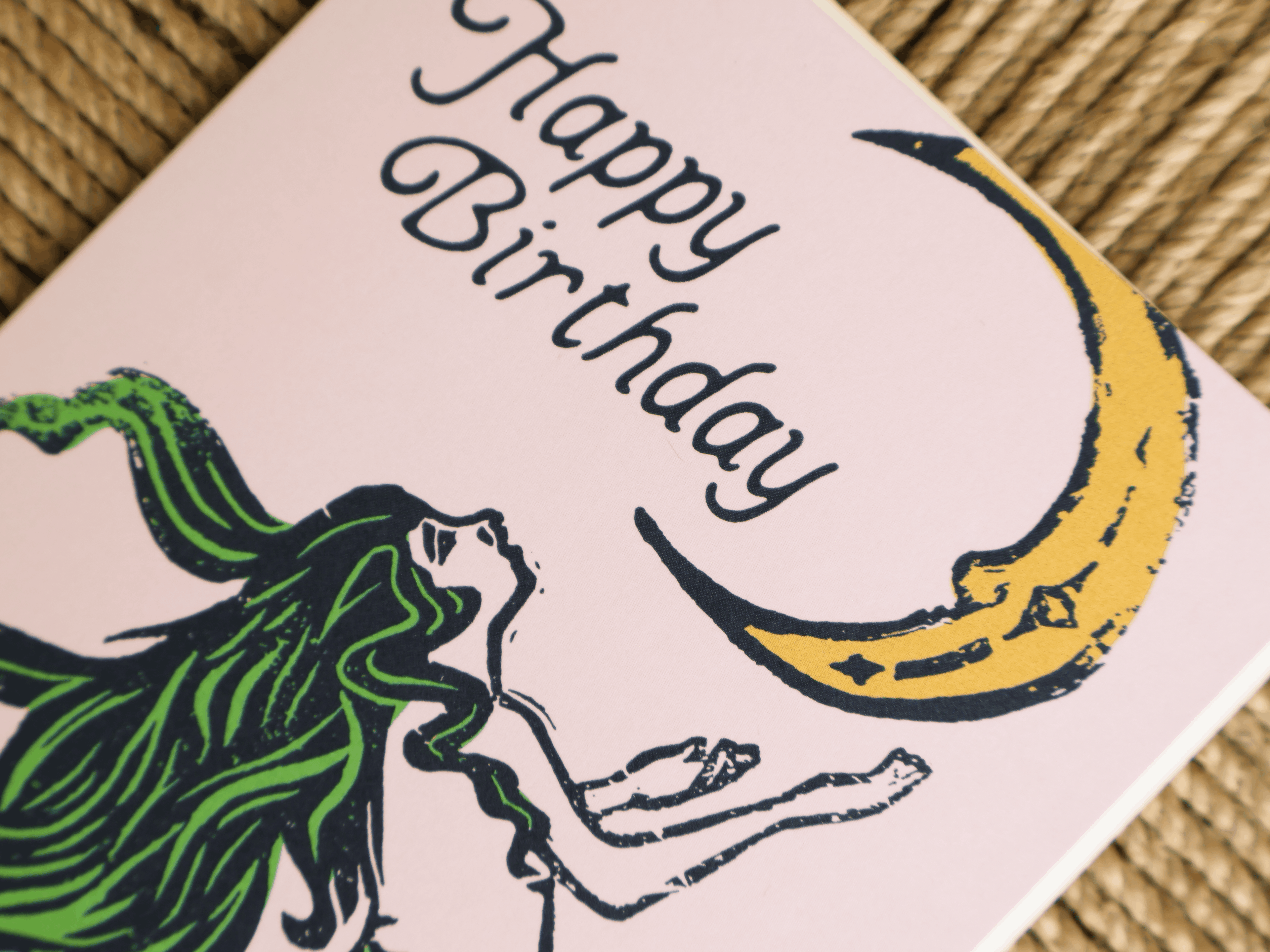 A close-up of a square greeting card with a pink background, featuring a stylized illustration of a woman with long green hair reaching toward a yellow crescent moon, with the words 'Happy Birthday' written in black script.