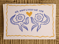 A square white greeting card with a hand-drawn blue and yellow design, featuring the text 'OH, HOW LUCKY WE ARE' above two stylized sun-like figures facing each other and a yellow heart in the center, placed on a woven brown surface.