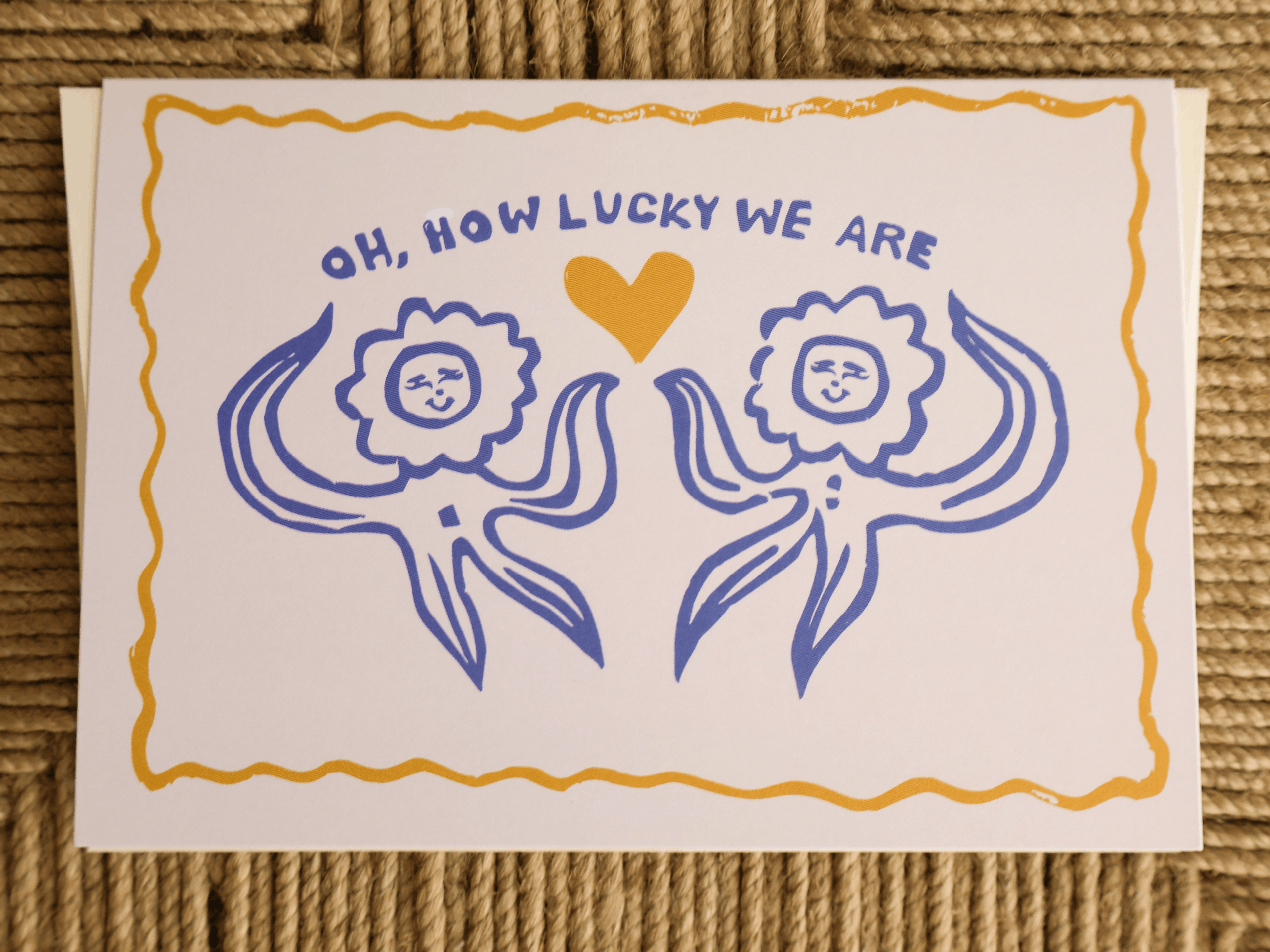 A square white greeting card with a hand-drawn blue and yellow design, featuring the text 'OH, HOW LUCKY WE ARE' above two stylized sun-like figures facing each other and a yellow heart in the center, placed on a woven brown surface.