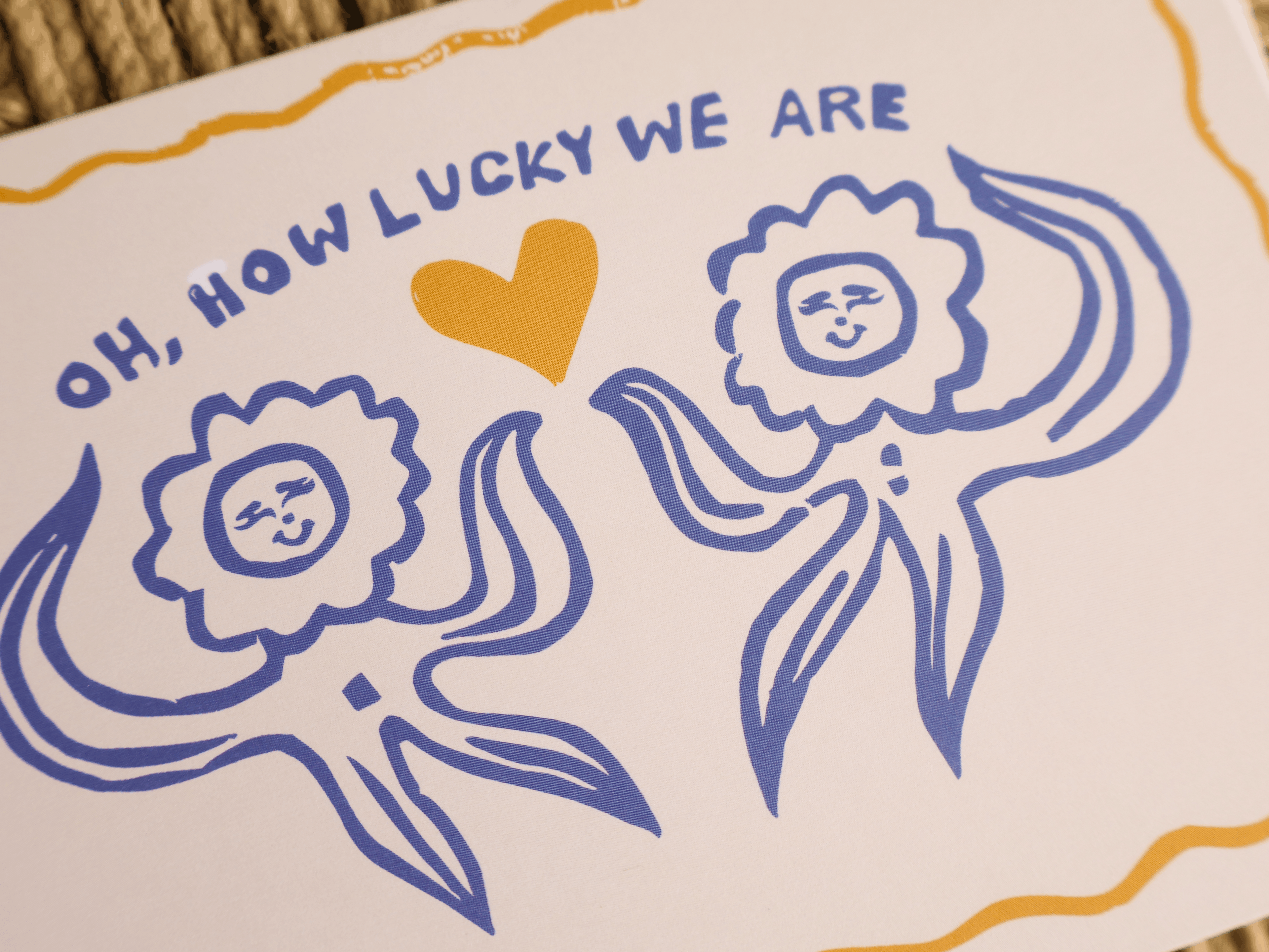 A close-up of a printed card or paper featuring a hand-drawn style illustration with the text 'OH, HOW LUCKY WE ARE' above two blue sun-like figures and a small yellow heart on an off-white background.
