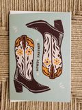 A square greeting card featuring a western-themed illustration of two cowboy boots with the text 'HOWDY, PARTNER' on a light sage green background, placed on a woven straw surface.