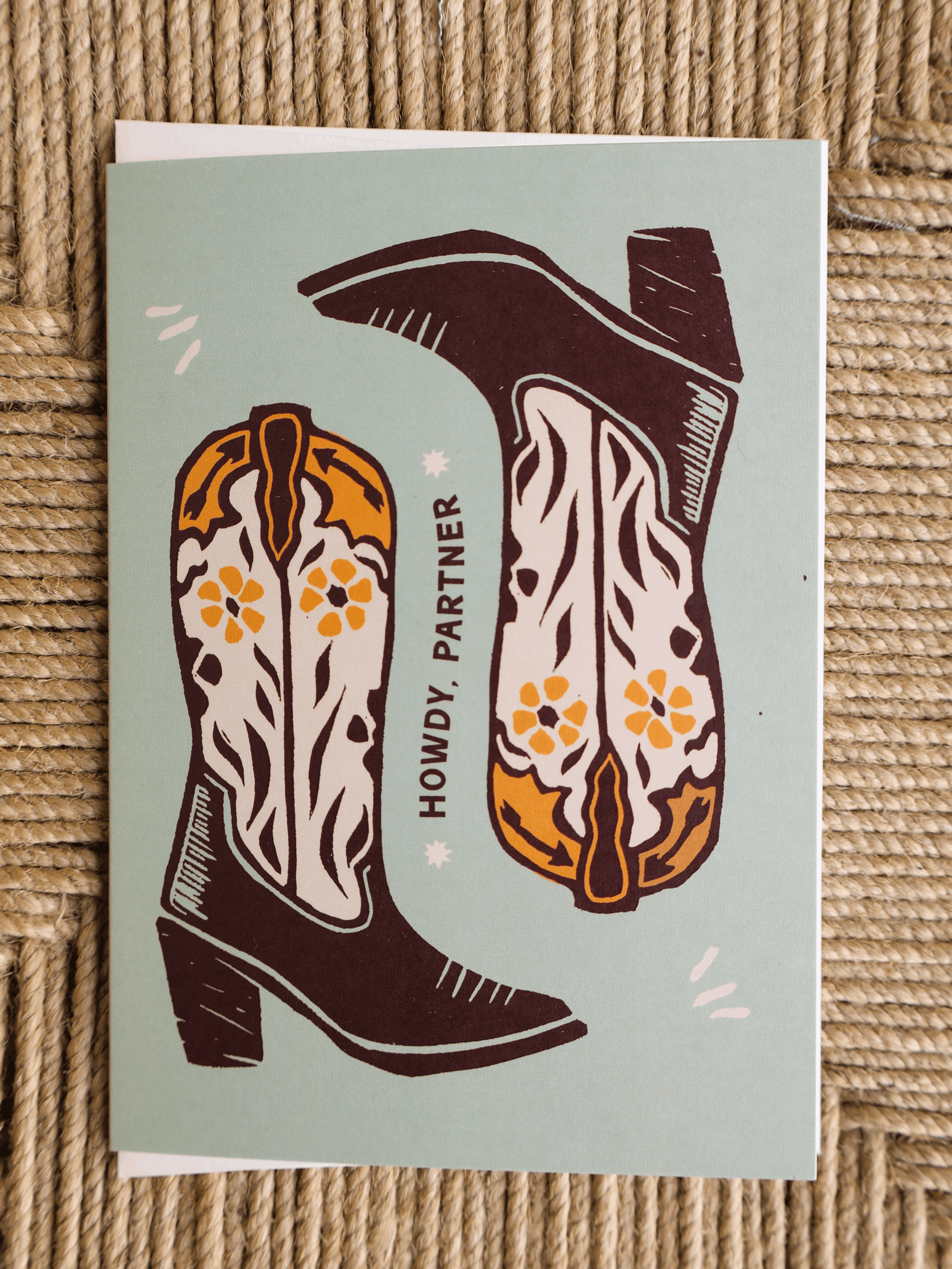 A square greeting card featuring a western-themed illustration of two cowboy boots with the text 'HOWDY, PARTNER' on a light sage green background, placed on a woven straw surface.