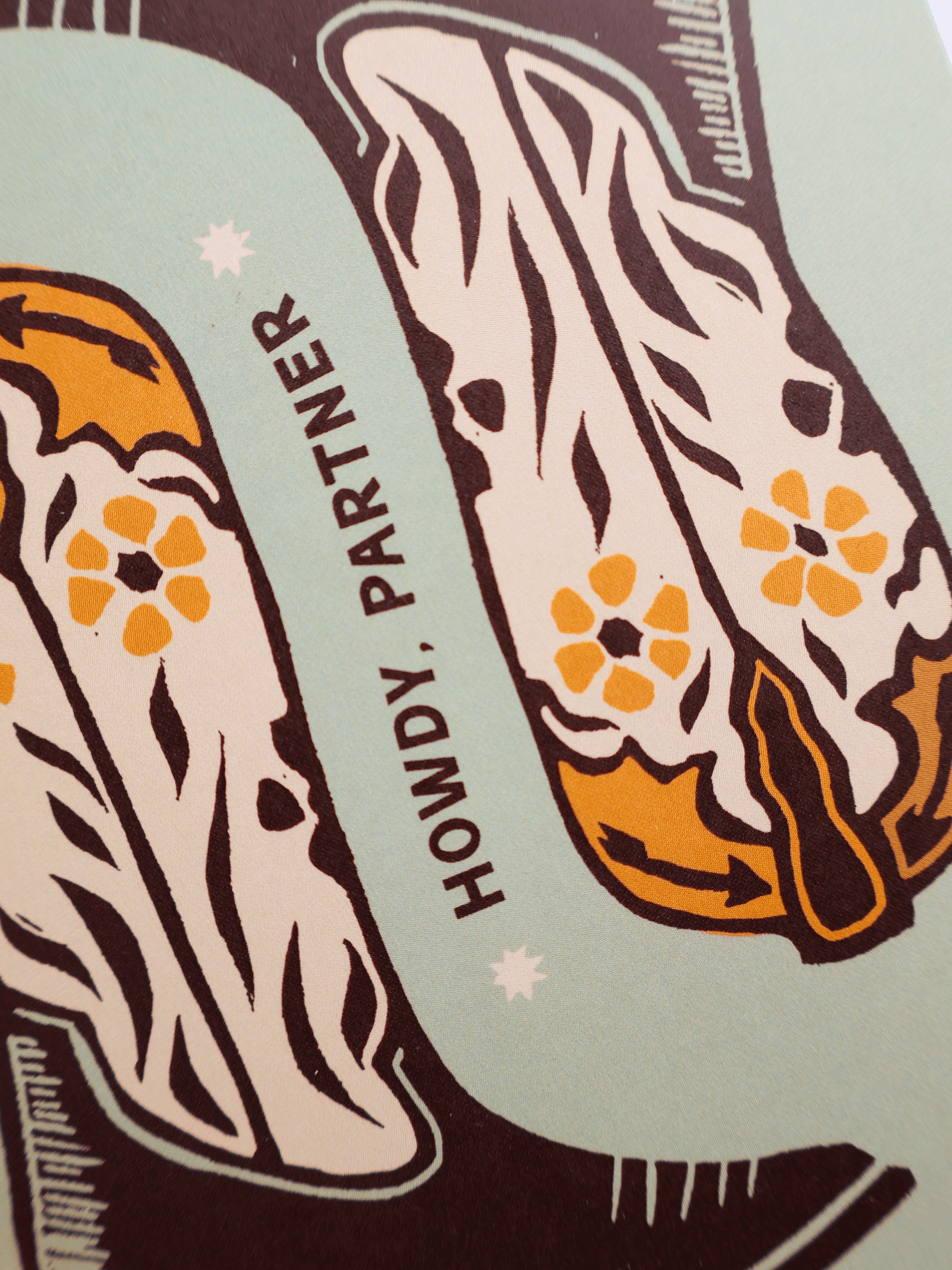 A close-up of a graphic print featuring two stylized cowboy boots in cream, orange, and dark brown, with the text 'HOWDY, PARTNER' printed vertically between them on a pale mint background.