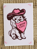 A square greeting card featuring a cartoon dog wearing a pink cowboy hat and holding a pink heart that says 'I WOOF YOU', photographed on a woven beige surface.