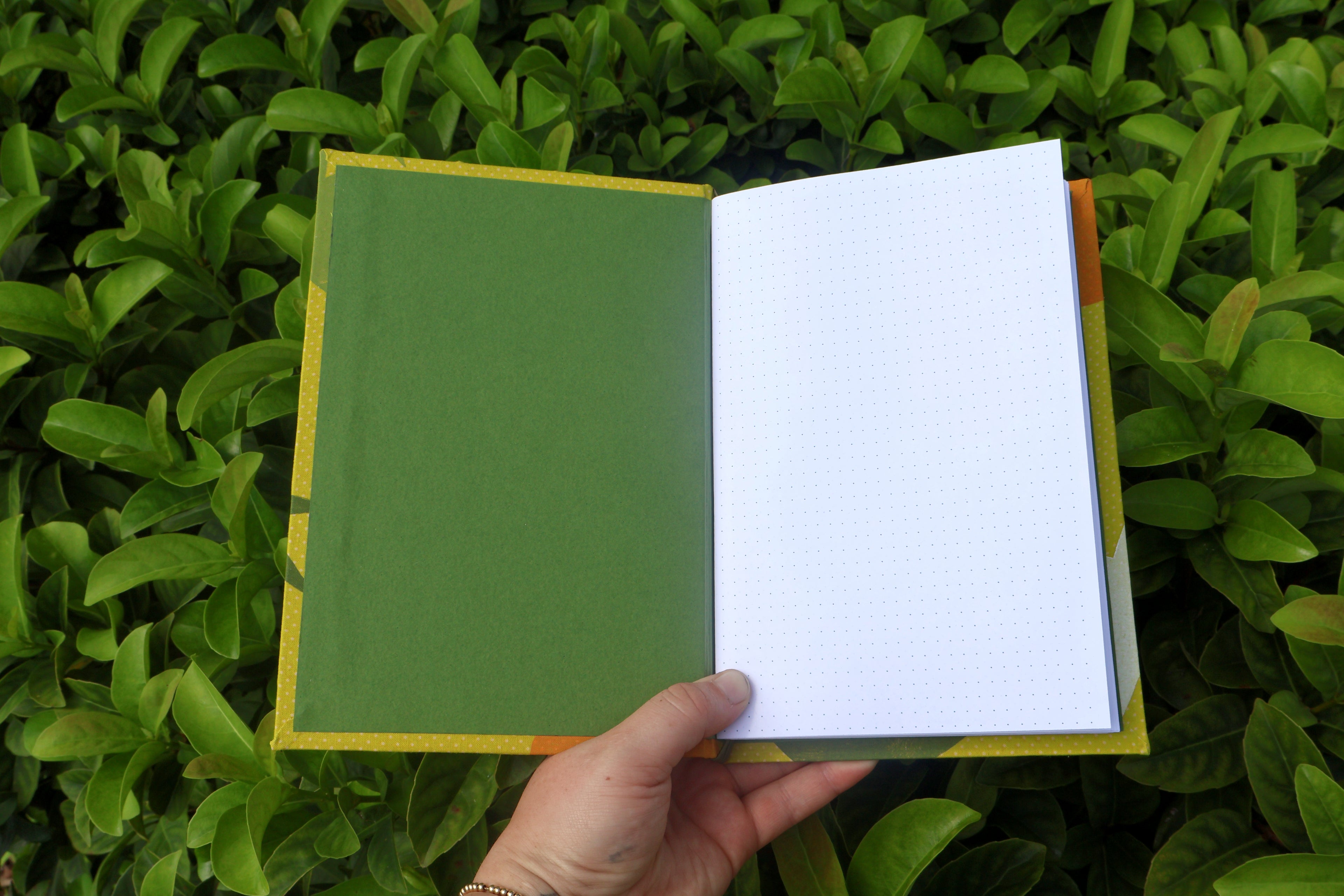 A hand holds opened yellow-patterned refillable bullet journals in front of dense green foliage. green interior cover paper and white bullet journal paper. hand bound and linocut printed with more love studio