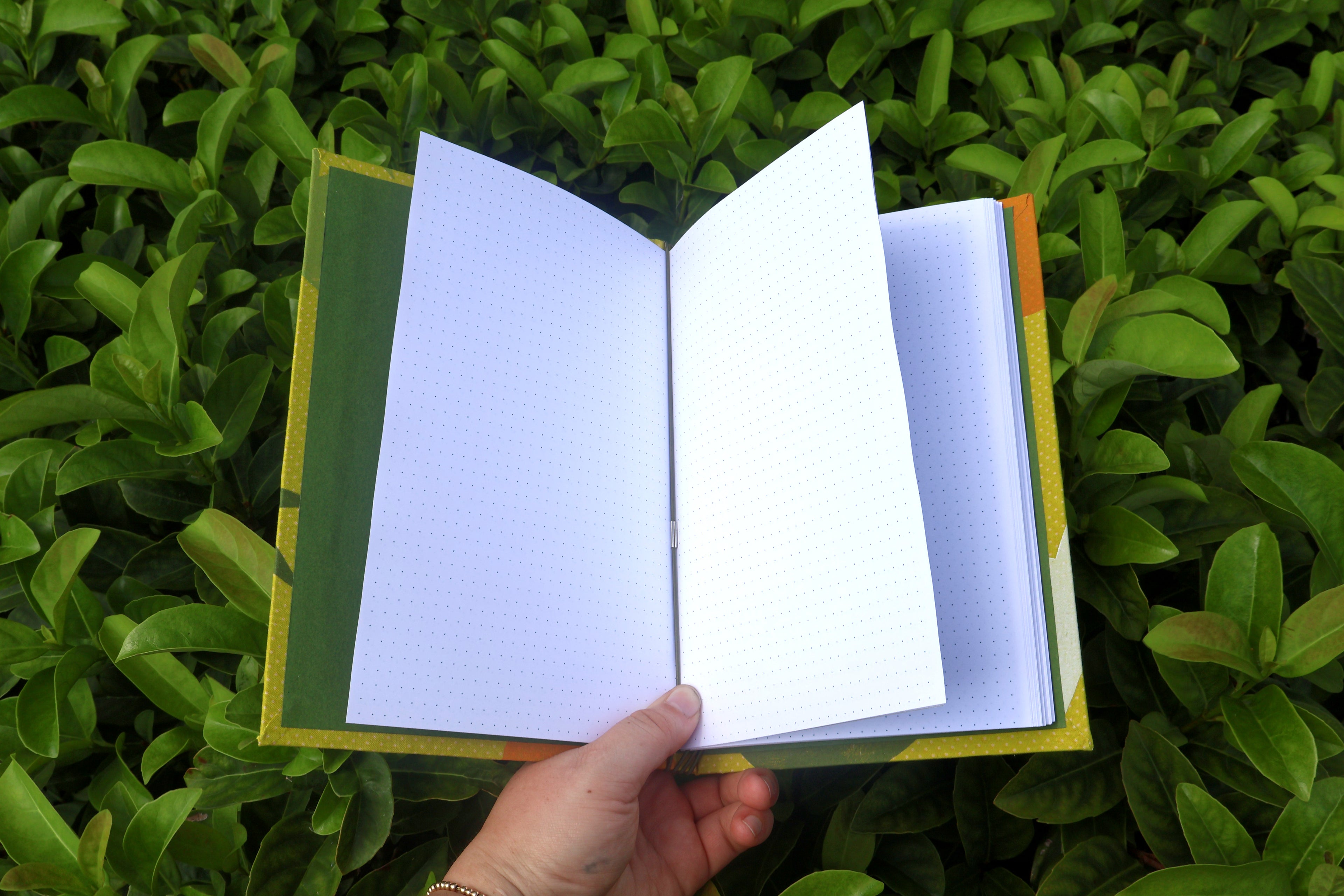 An open notebook with white bullet journal pages held by a hand against a background of green leaves. hand bound and linocut printed with more love studio