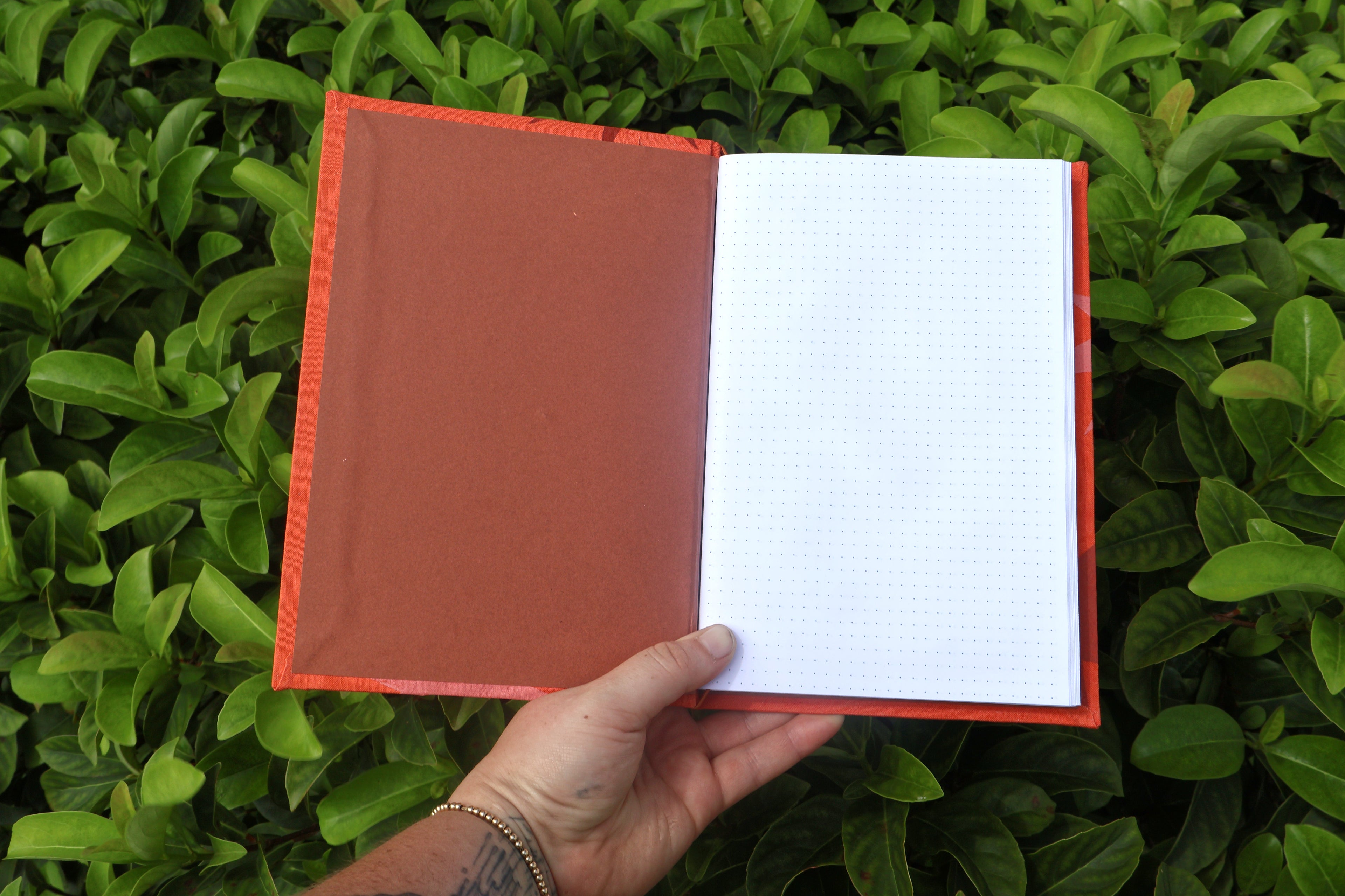An open refillable journal with a reddish-brown cover and white graph paper pages, held by a hand against a background of green leaves. hand bound and linocut printed with more love studio
