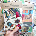 Dovetail Junk Journaling Kit held by a hand with various items including stickers, ribbon, and decorative paper.