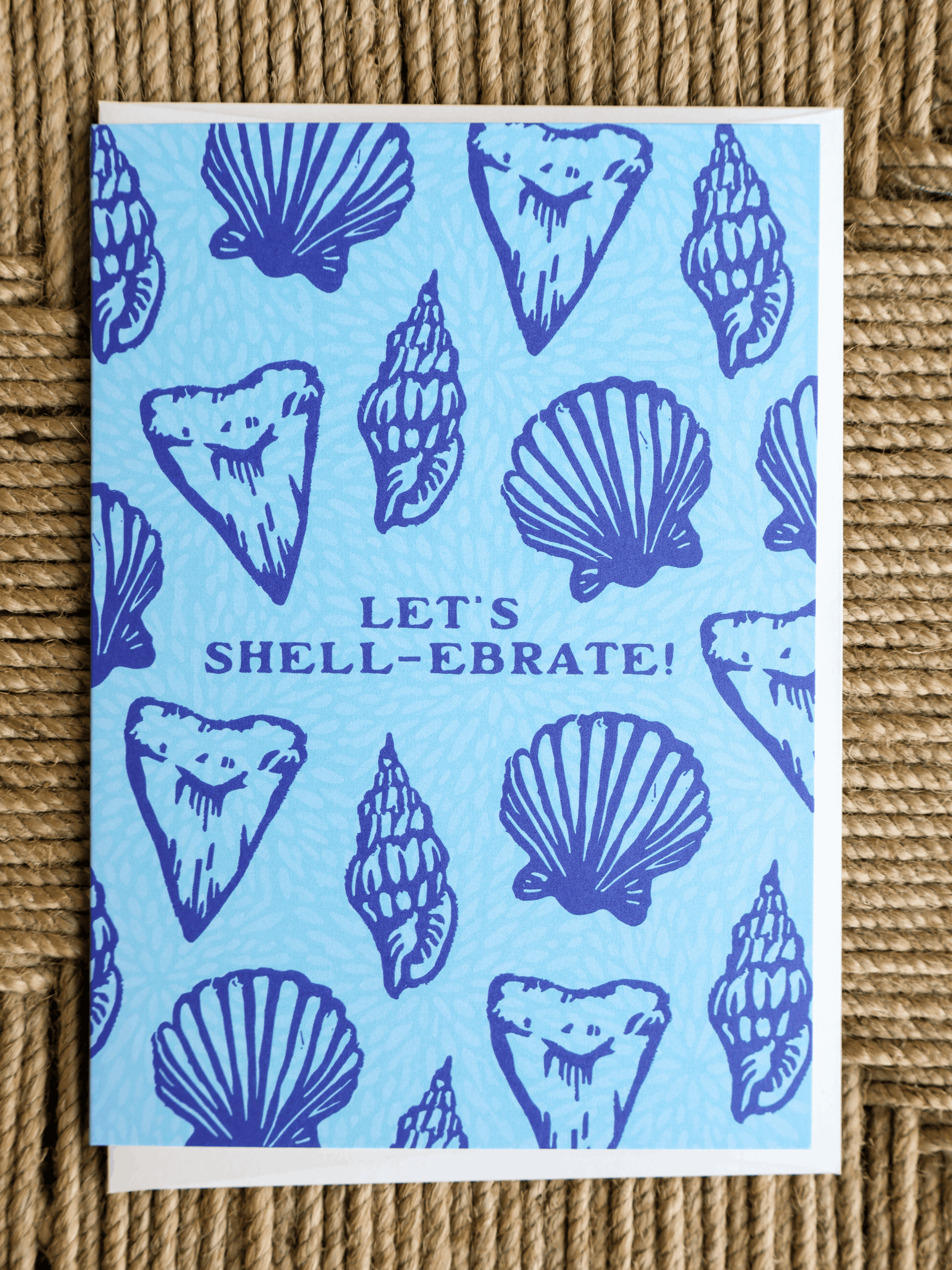 A square greeting card featuring a light blue background with dark blue illustrations of seashells and the text 'LET'S SHELL-EBRATE!' in the center, photographed on a woven straw surface.