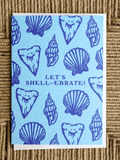 A square greeting card featuring a light blue background with dark blue illustrations of seashells and the text 'LET'S SHELL-EBRATE!' in the center, photographed on a woven straw surface.