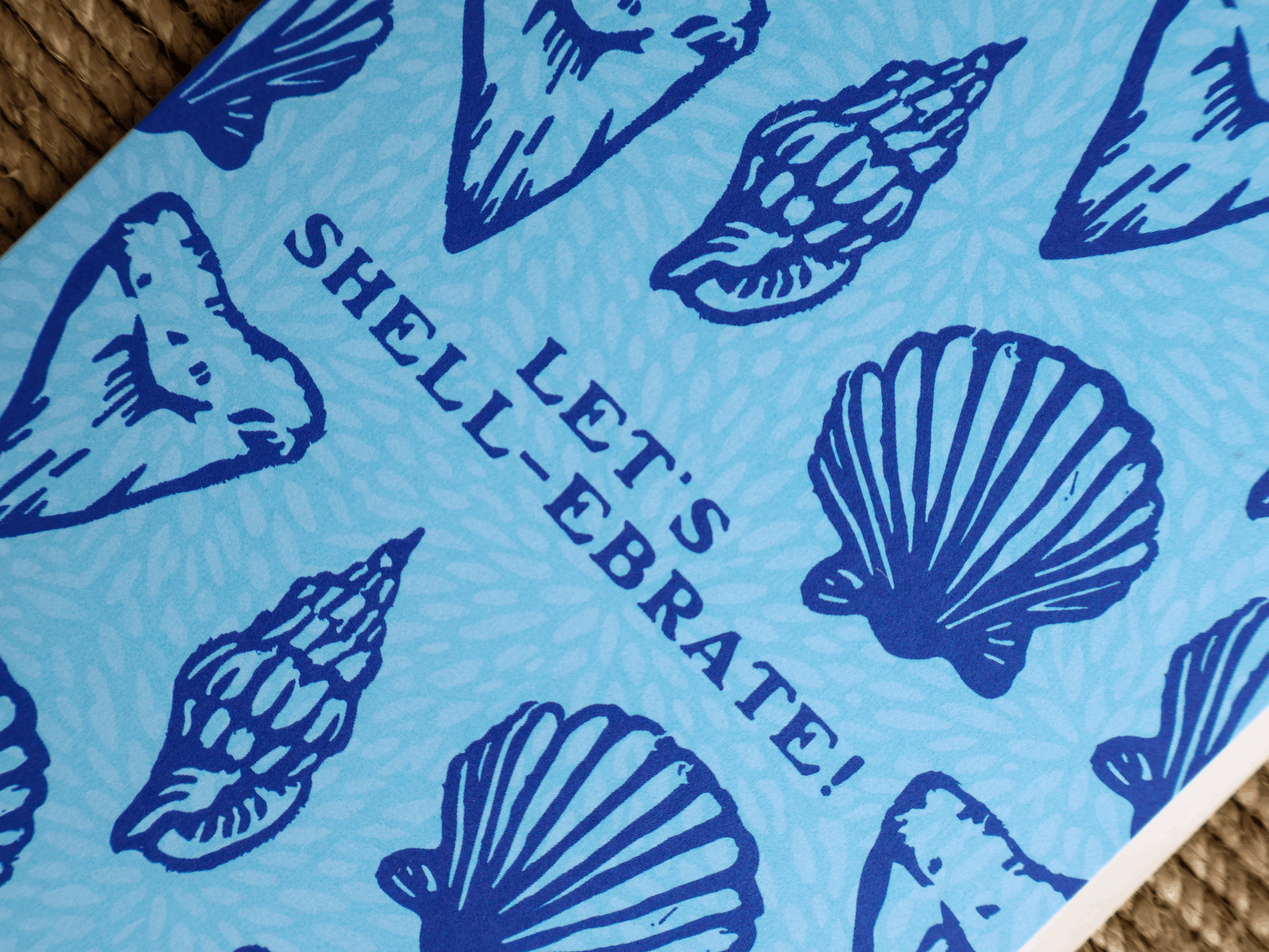 A close-up of a blue patterned card or paper featuring navy shell illustrations and the text 'LET'S SHELL-EBRATE!' on a woven brown surface.
