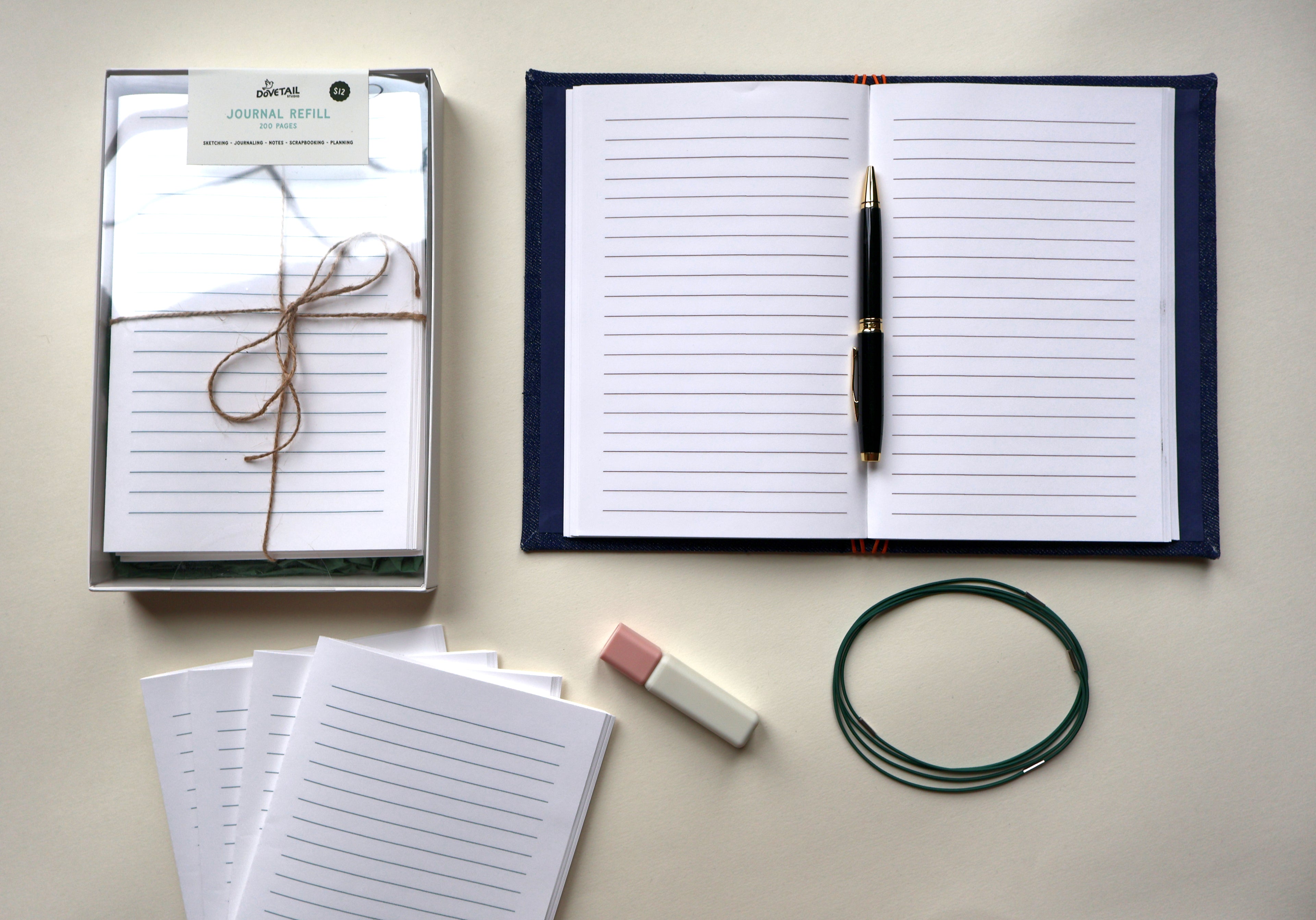 A styled flat-lay of stationery items on a light beige surface: an open navy-bound lined journal with a black and gold pen, a boxed pack of lined paper tied with twine, several loose lined sheets, a pink-and-white highlighter, and a dark green loop of elastic journal binders. Showcasing Dovetail Studio's handmade refillable journal refills