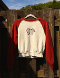A red and off-white raglan crewneck sweatshirt hanging on a wooden fence outdoors. The front features a black graphic of two cartoon characters with a speech bubble that reads 'you're cute'.