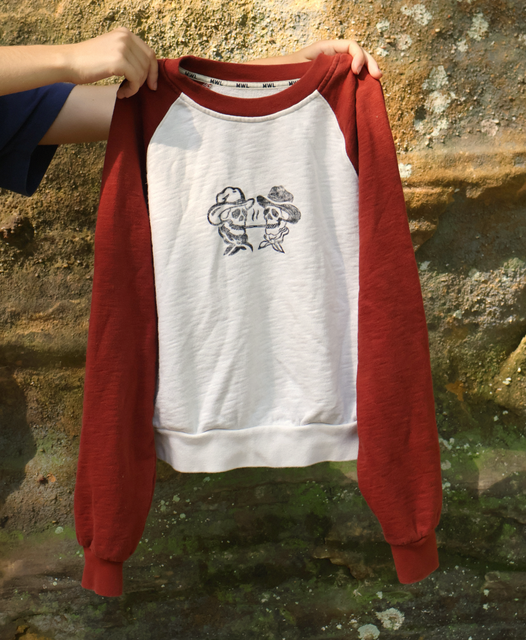 A person holds up a raglan crewneck sweatshirt with a white body and dark red sleeves, featuring a black graphic of two cartoon characters wearing hats on the chest. The background is a textured, mossy stone wall.