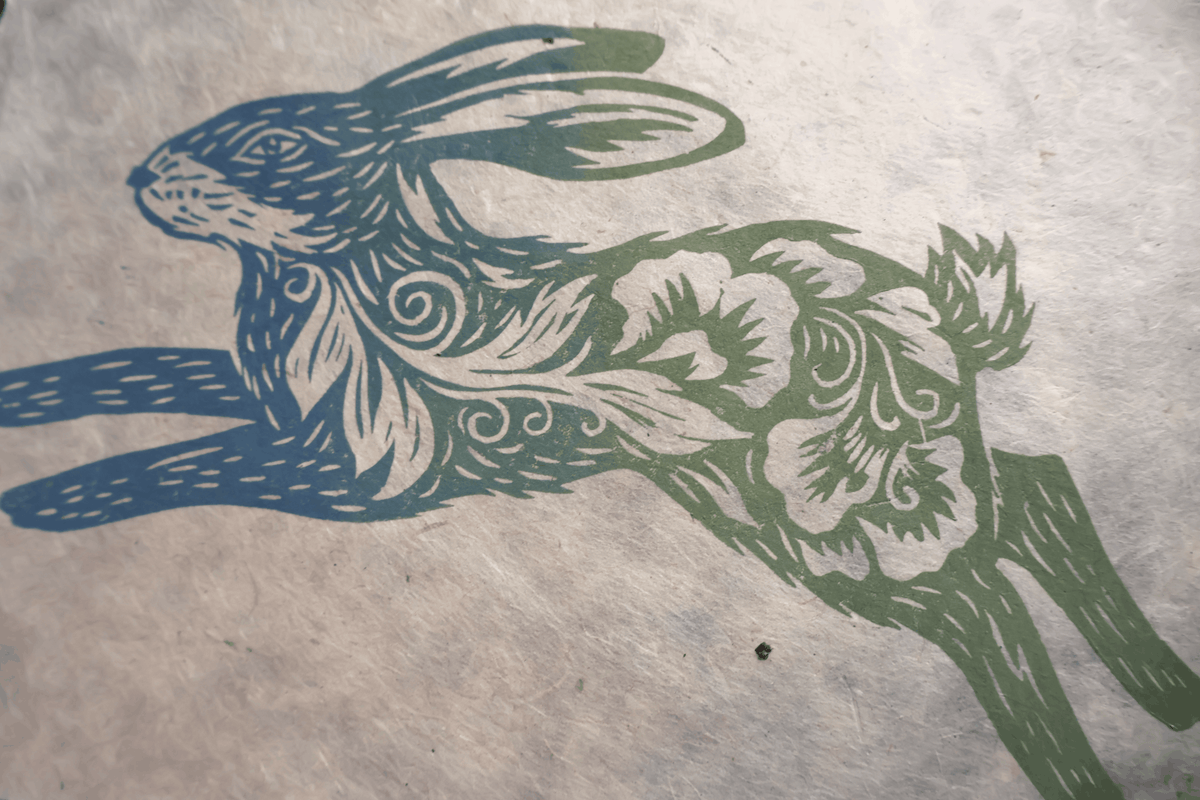 A stylized rabbit illustration printed on textured paper, with blue and green linocut-style patterns over a light beige background.