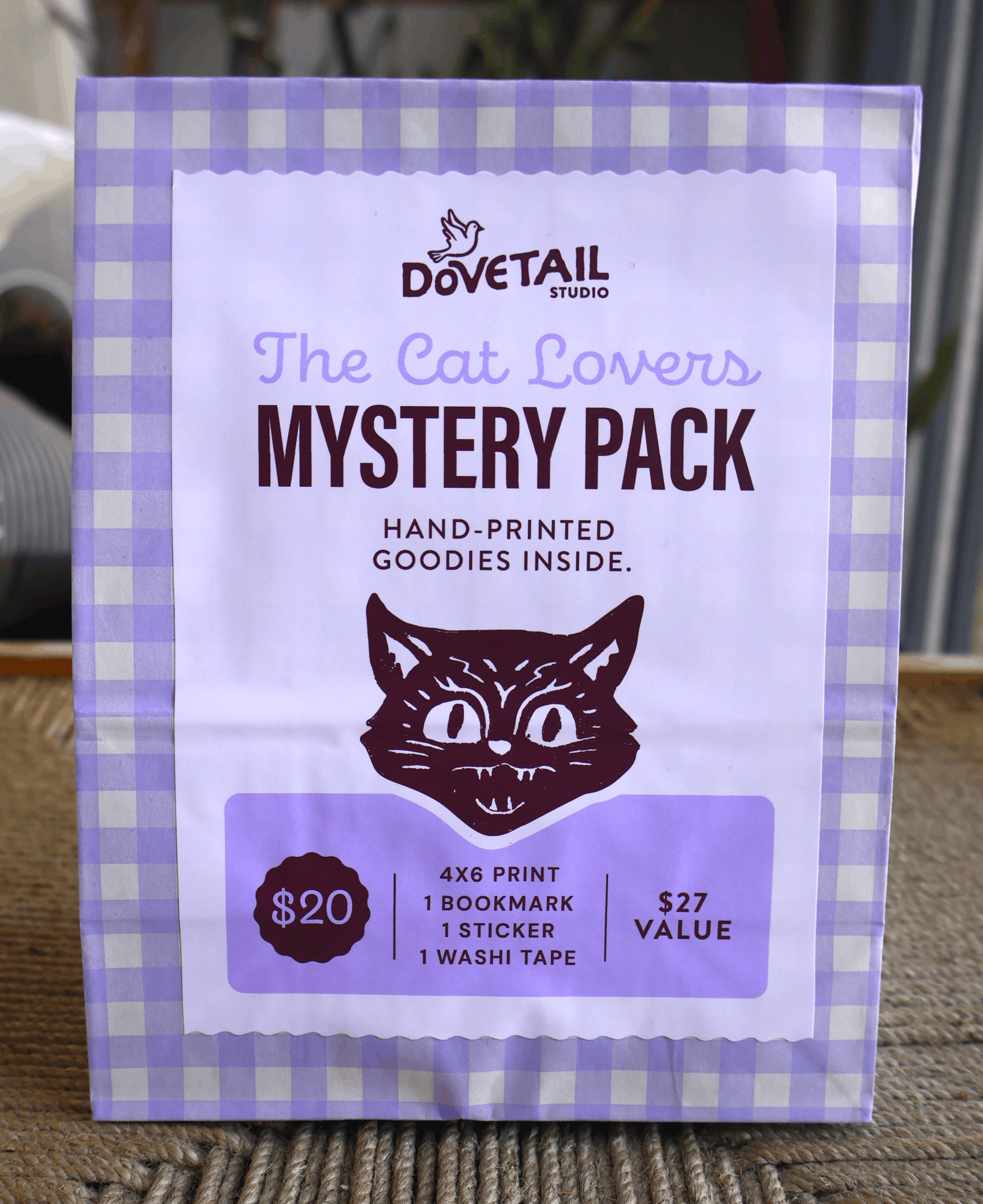 A package of Dovetail Studio 'The Cat Lovers Mystery Pack' featuring a lavender and white gingham border, a white center label with a dark purple cat illustration, and product details including a $20 price and $27 value.