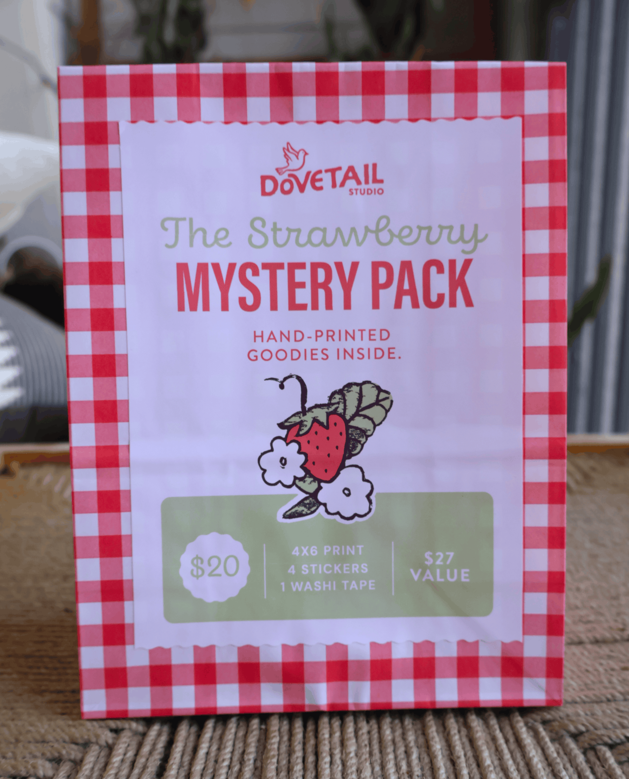 A package for a 'Dovetail Studio' Strawberry Mystery Pack, featuring a red and white gingham border and a white center with text and a strawberry sticker illustration, standing on a woven mat.