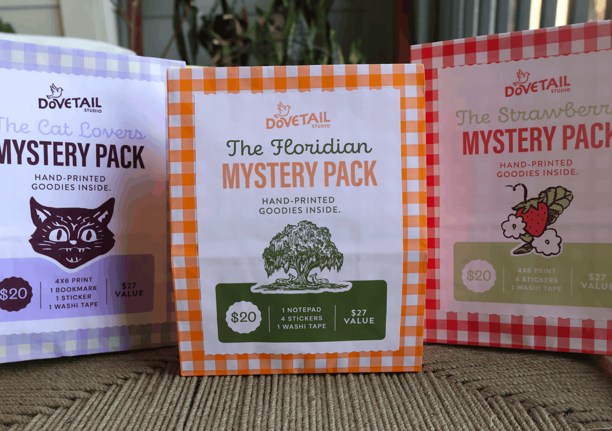 Three boxed pet treats packages from Dovetail Products are lined up on a woven surface. The center package is for 'The Floridian Mystery Pack' with an orange-and-white gingham border, and the packages on the left and right show 'The Cat Lover Mystery Pack' and 'The Strawberry Mystery Pack' with lavender and red gingham borders.