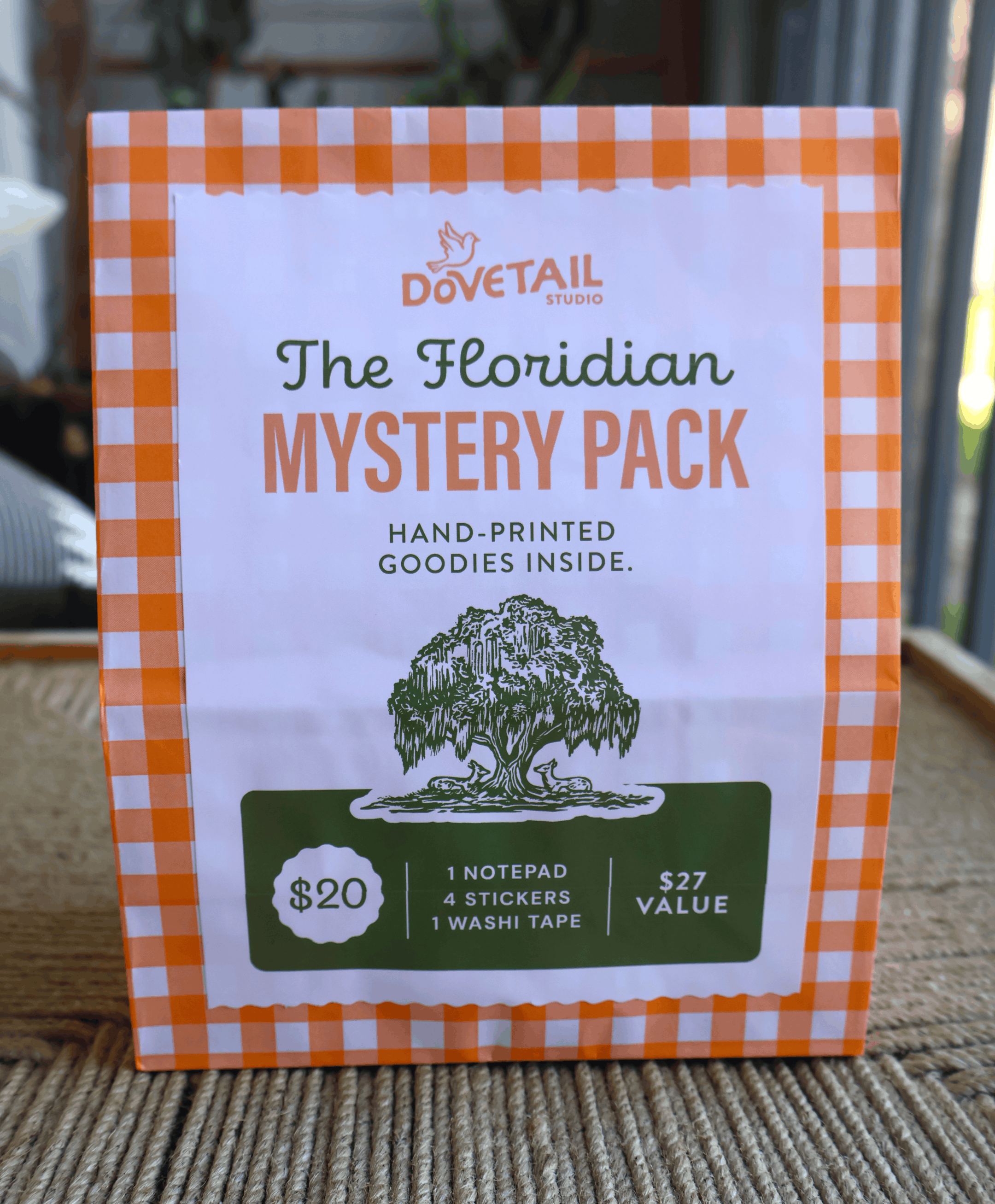 A boxed product labeled 'The Floridian Mystery Pack' by Dovetail Studio, standing on a woven surface. The package has an orange-and-white gingham border, a white center panel with green and orange text, and a dark green label near the bottom.