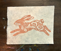 A rectangle piece of textured handmade paper featuring a printed rabbit illustration in terracotta orange on a warm off-white background, placed on a dark wooden surface.