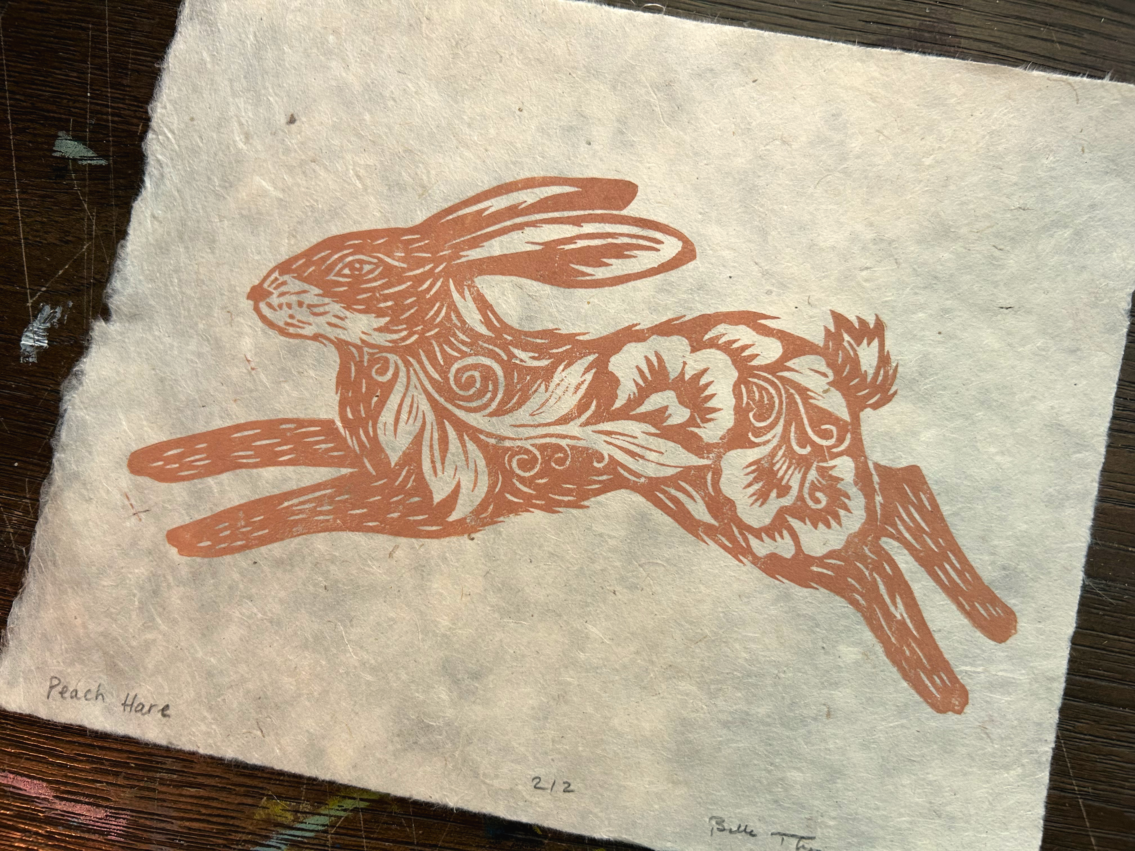 A square piece of textured handmade paper featuring a reddish-brown linocut or woodcut-style illustration of a leaping rabbit with floral details. Handwritten text at the bottom reads 'Peach Hare' and '2/2'.