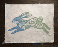 A square handmade paper print featuring a stylized rabbit cutout design in blue and green ink, placed on a dark wooden surface.