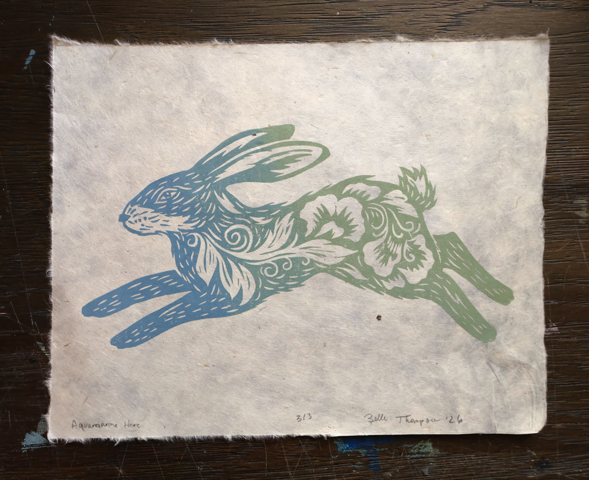 A square handmade paper print featuring a stylized rabbit cutout design in blue and green ink, placed on a dark wooden surface.