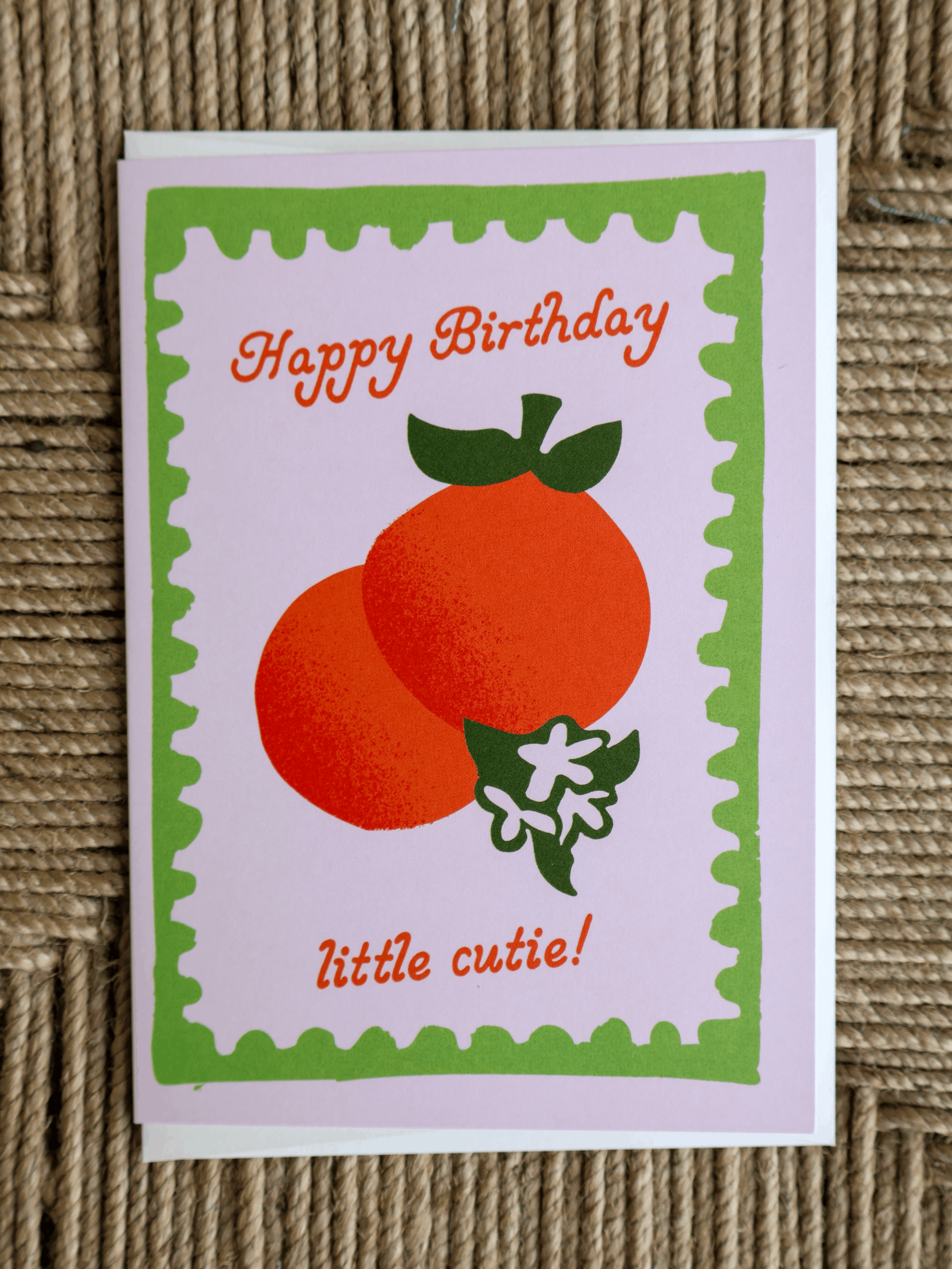 A square greeting card with a white background and a green scalloped border, featuring two orange fruits with green leaves and the text 'Happy Birthday little cutie!' in orange. The card is photographed on a textured brown woven surface.