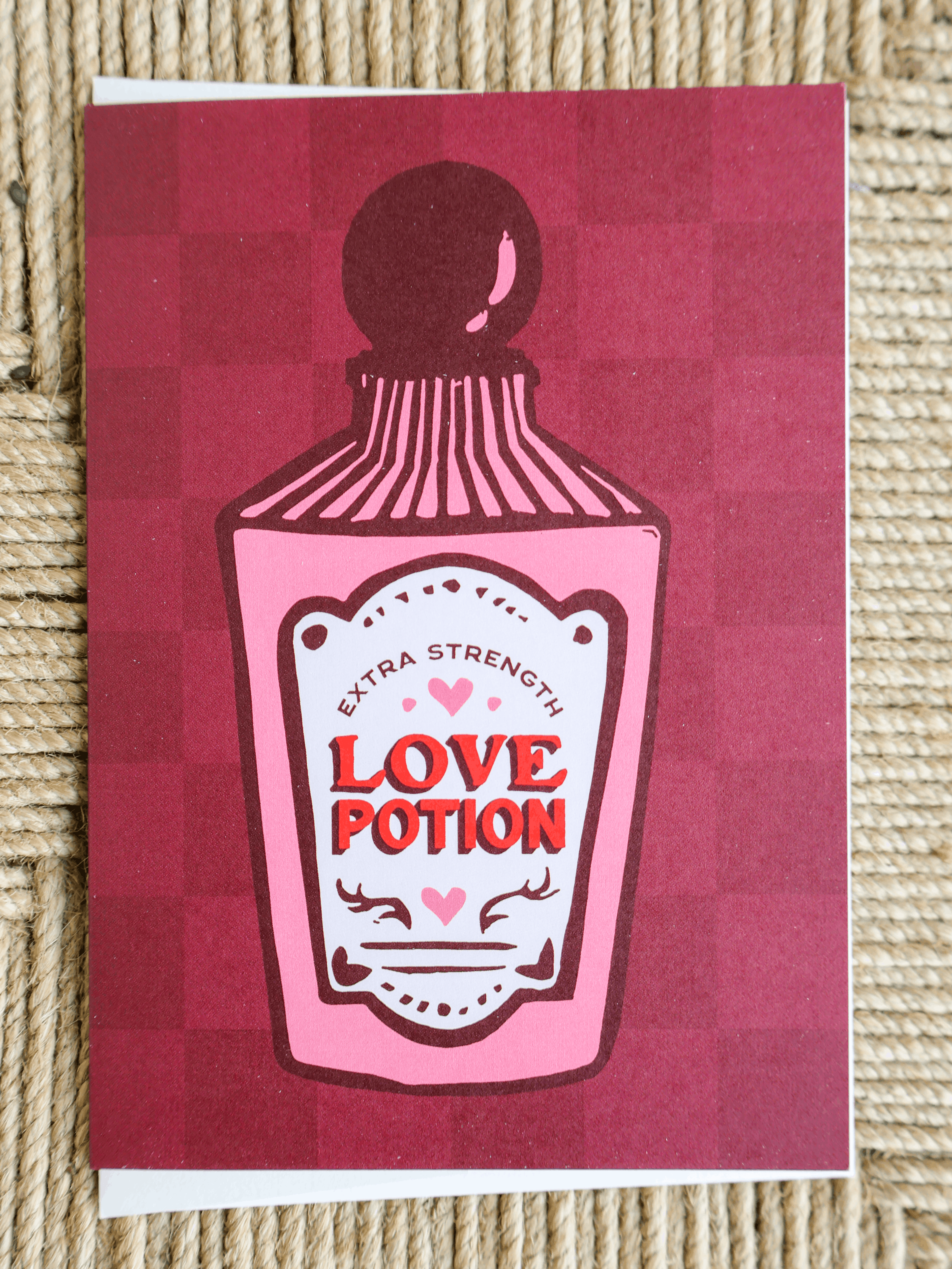 A square greeting card featuring a pink and red checkered background with a stylized illustration of a potion bottle labeled 'Extra Strength Love Potion'. The card is photographed flat on a textured beige woven surface.