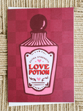 A square greeting card featuring a pink and red checkered background with a stylized illustration of a potion bottle labeled 'Extra Strength Love Potion'. The card is photographed flat on a textured beige woven surface.