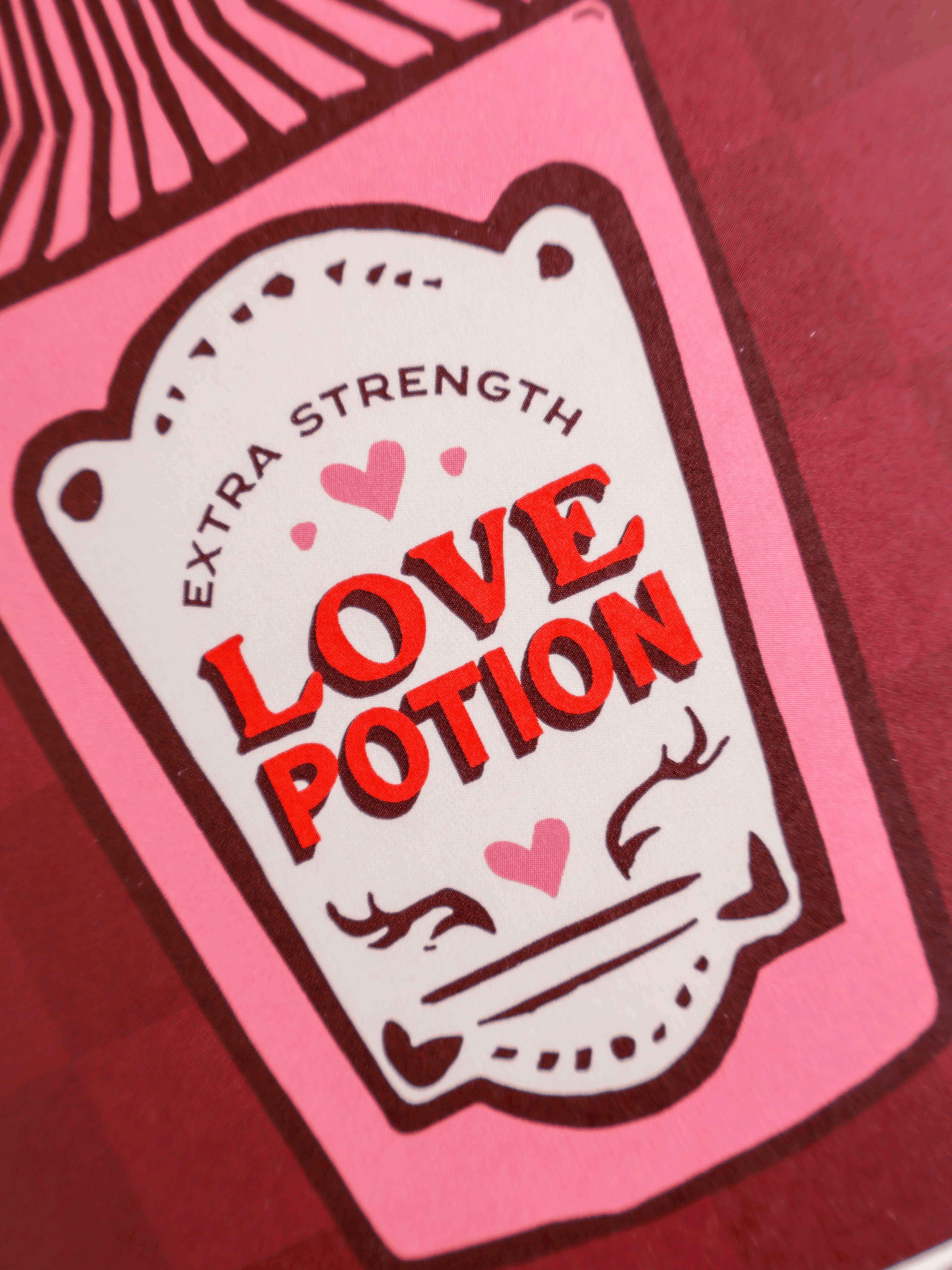 A close-up of a pink product package or label featuring the words 'EXTRA STRENGTH LOVE POTION' in red and black, with small pink hearts and illustrated faces on a red background.